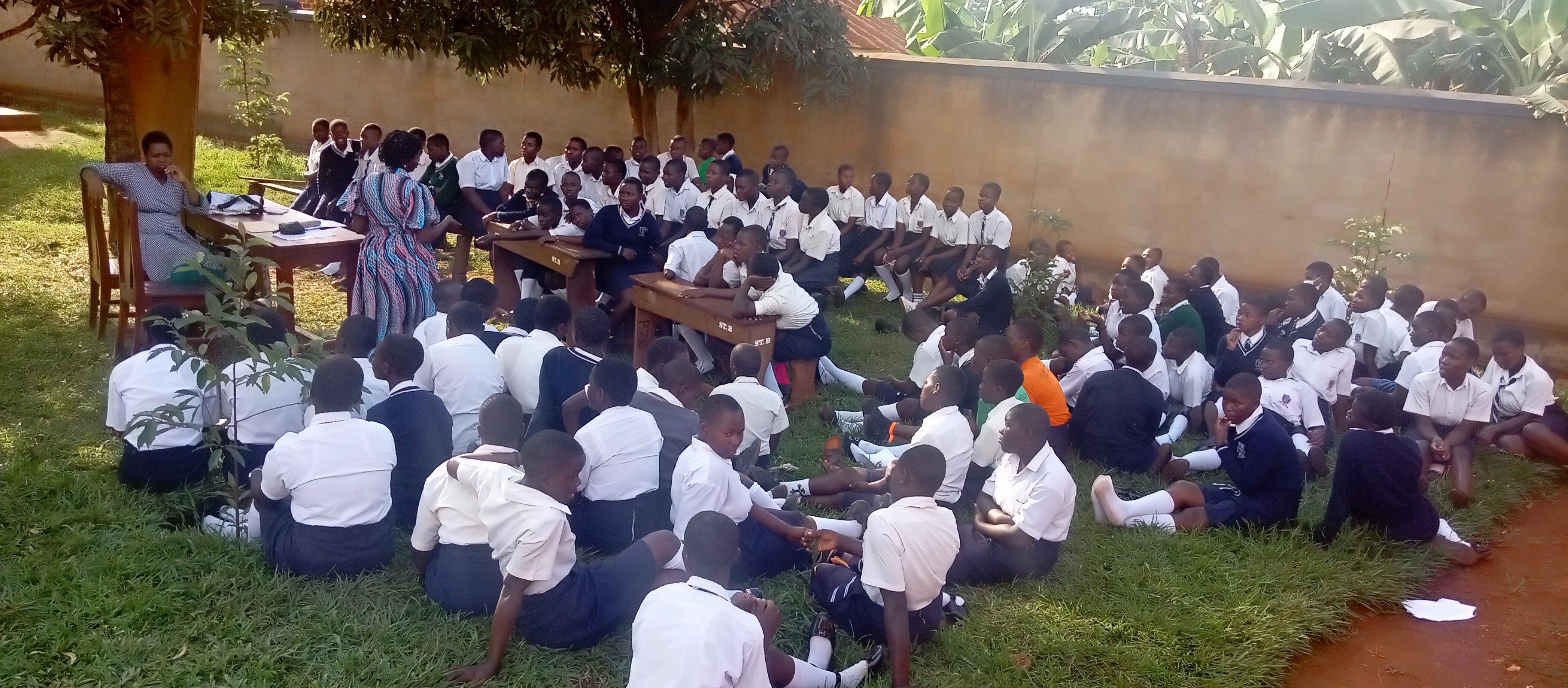 Students in uniforms sit outdoors on the grass listening to a speaker during a lesson