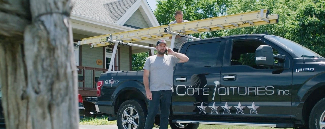Deux hommes en tenue de travail près d'une camionnette noire avec une échelle sur le dessus devant une maison résidentielle.