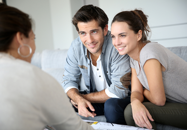 Un couple souriant discute avec une conseillère assise en face, autour de documents posés sur une table.