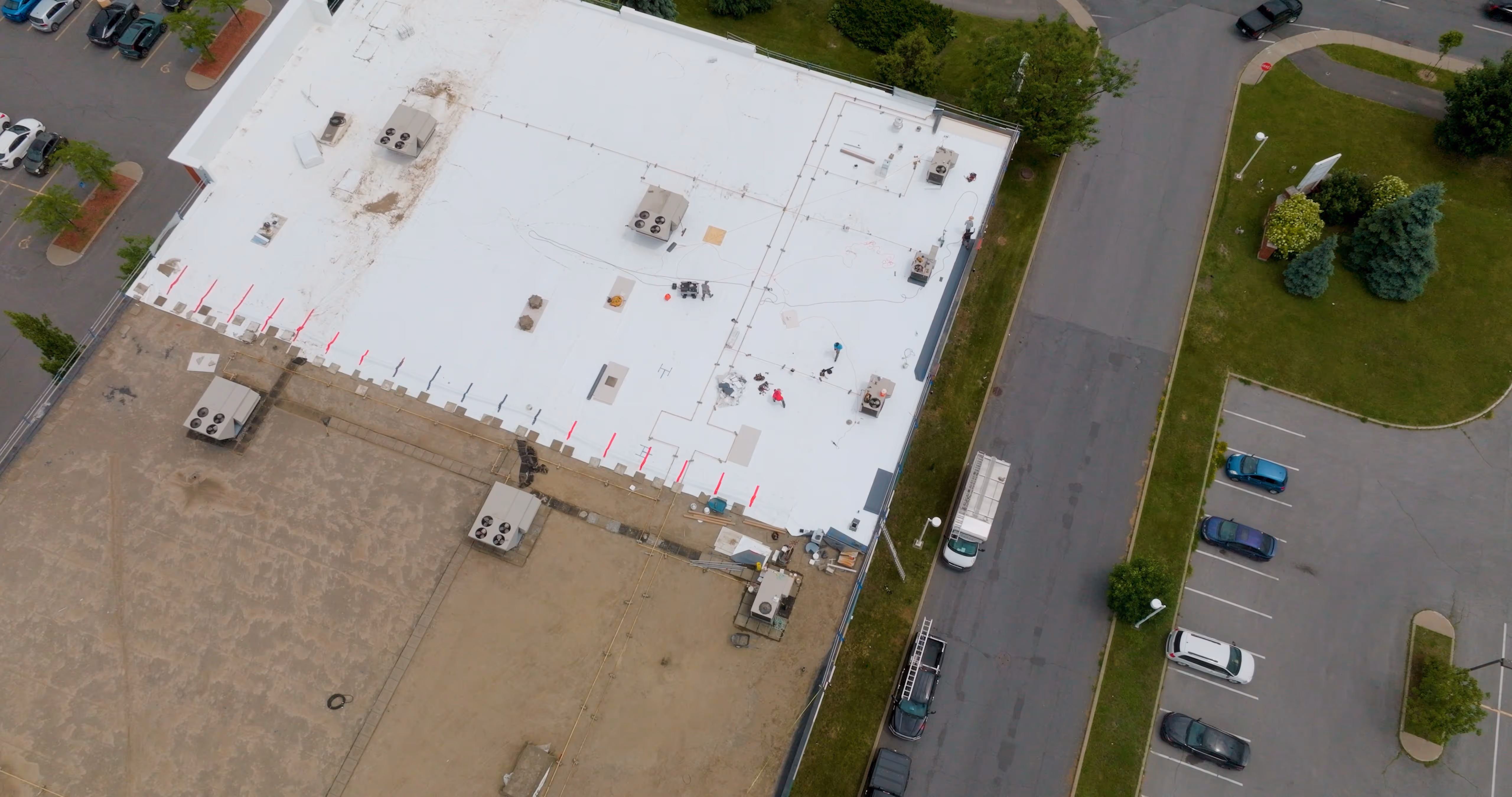 Vue aérienne montrant la moitié d'un toit de bâtiment en cours d'installation avec une surface blanche neuve et l'autre moitié encore à nu, avec des ventilateurs et travailleurs présents.