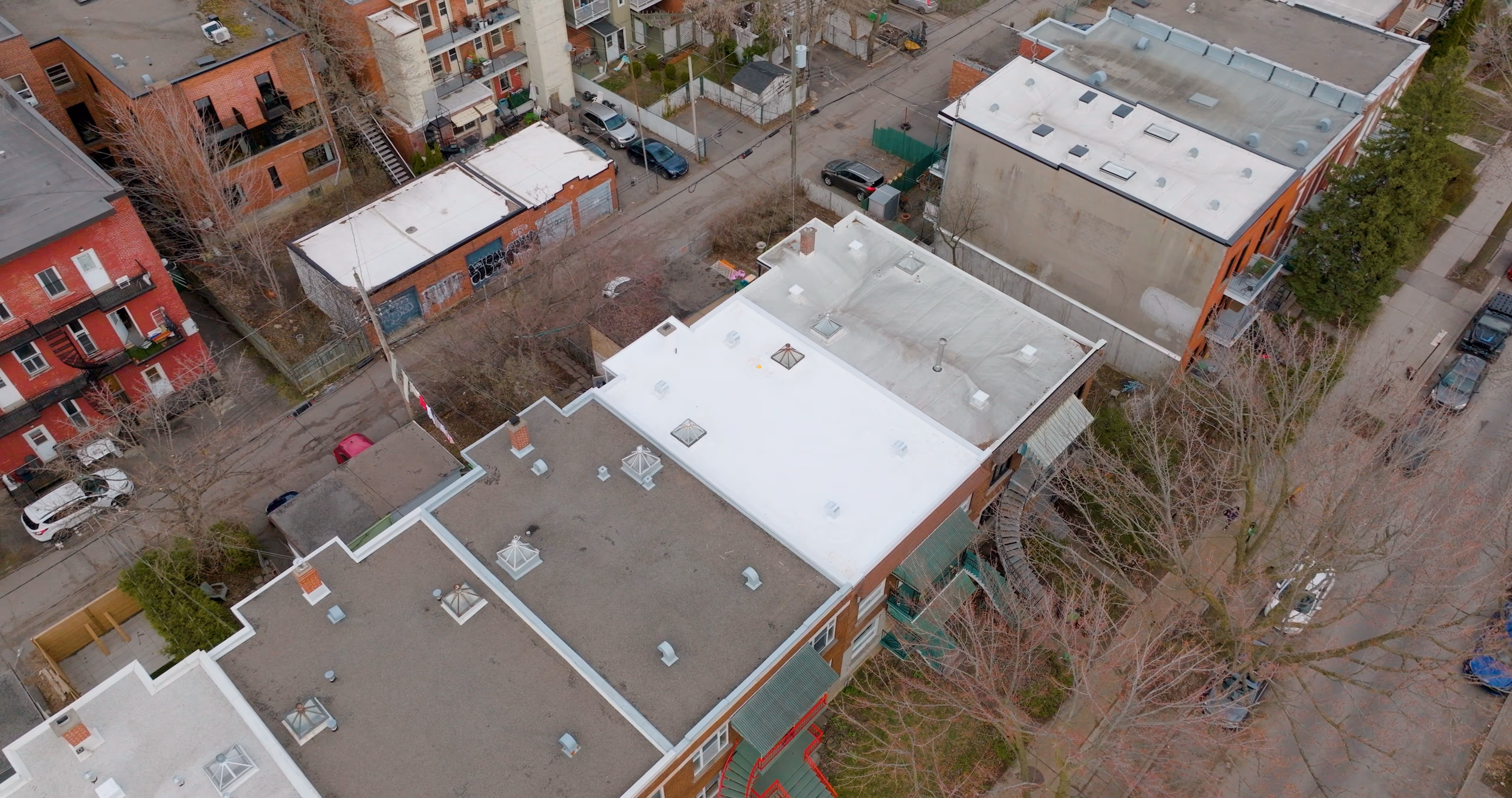 Vue aérienne des toits plats de plusieurs bâtiments résidentiels dans un quartier urbain, avec des voitures garées le long des rues et des arbres sans feuilles.