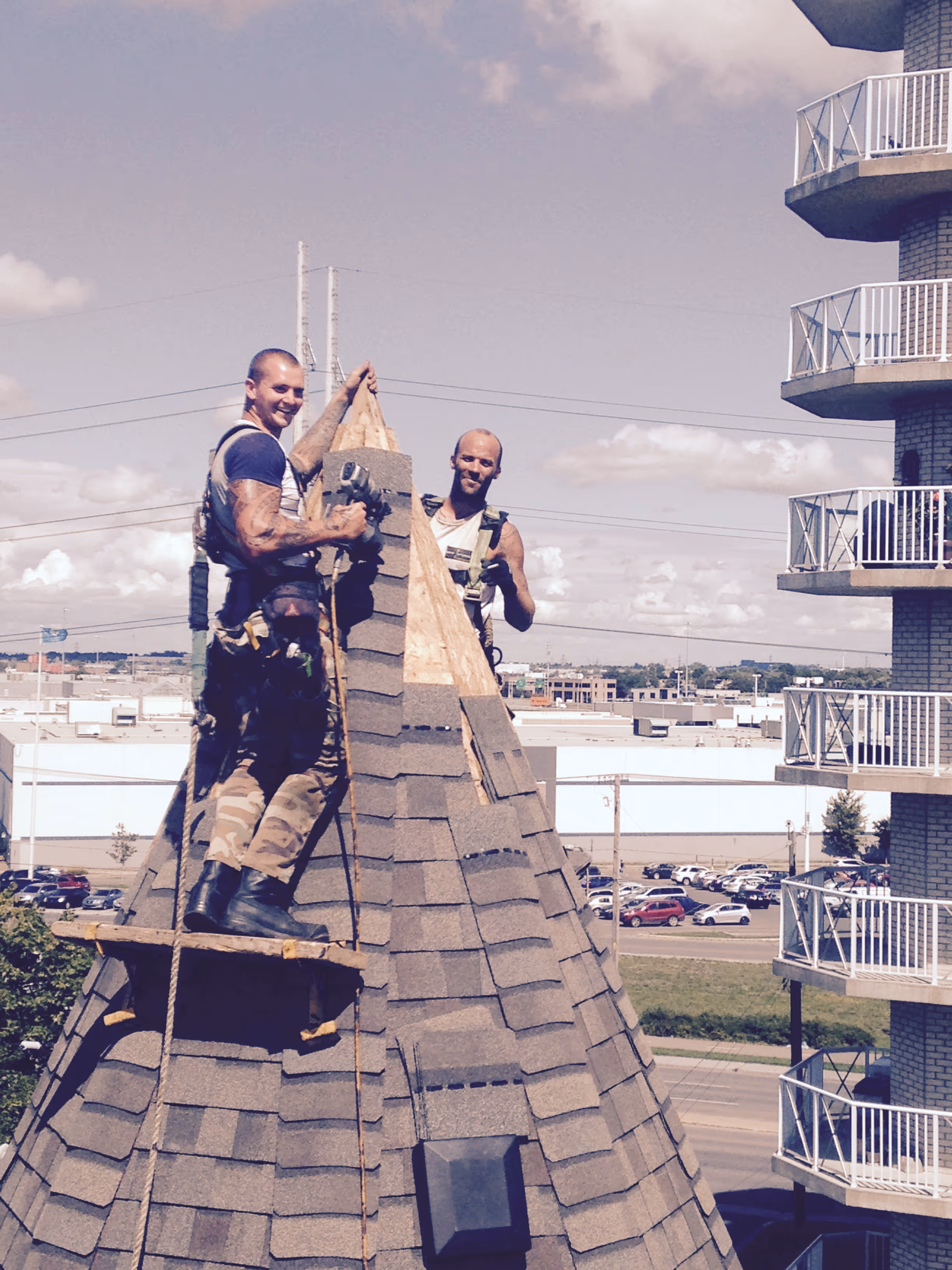 Deux ouvriers du bâtiment sur un toit conique en ardoises en train de poser le sommet sous un ciel partiellement nuageux.