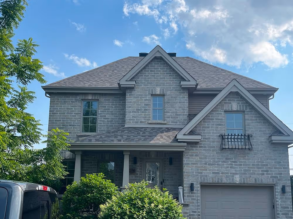 Maison moderne en brique grise avec un garage, une porte d'entrée sous un porche et des arbustes au premier plan sous un ciel bleu partiellement nuageux.