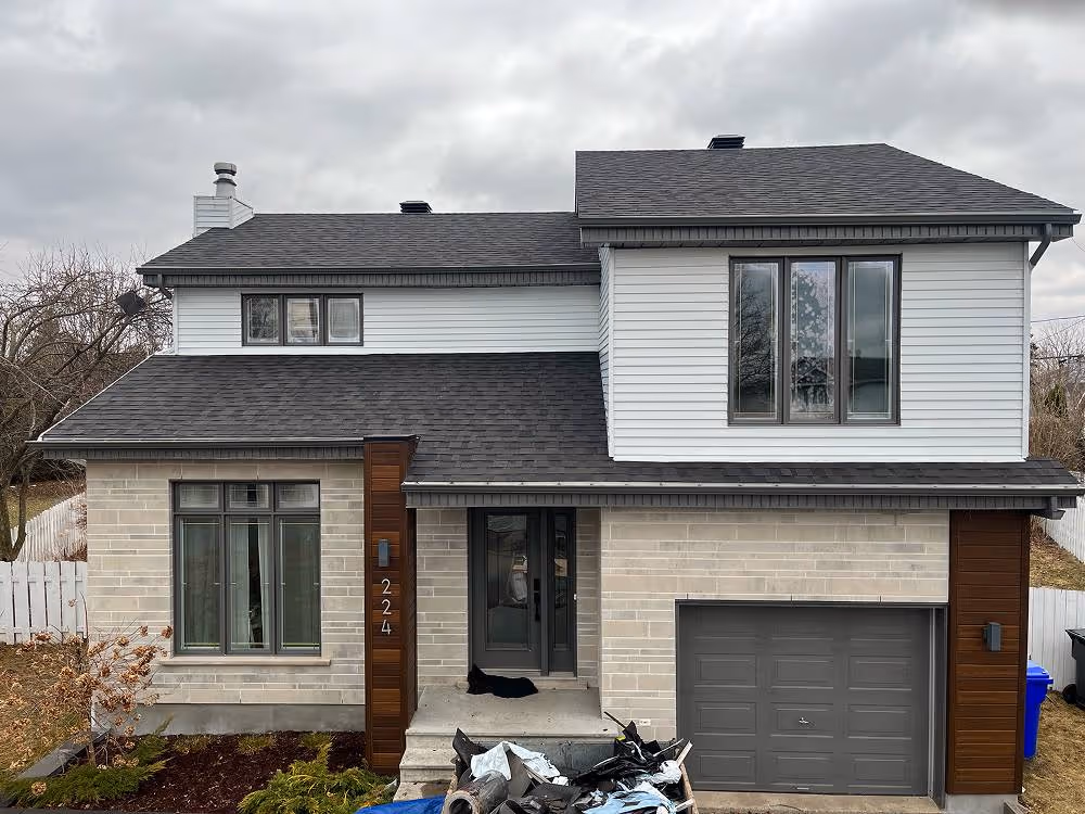 Maison moderne à deux étages avec façade en briques claires, un garage gris, et un chat noir allongé sur le seuil d'entrée.