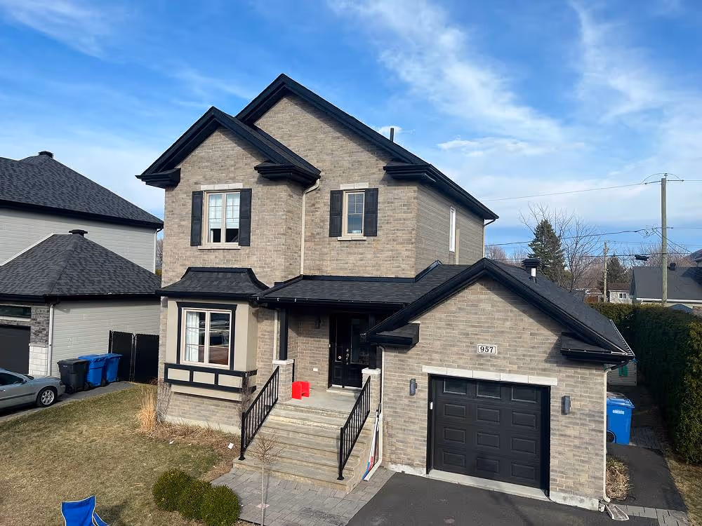 Maison moderne en briques grises avec un garage noir, une porte d'entrée double noire et un escalier menant à la porte principale sous un ciel partiellement nuageux.