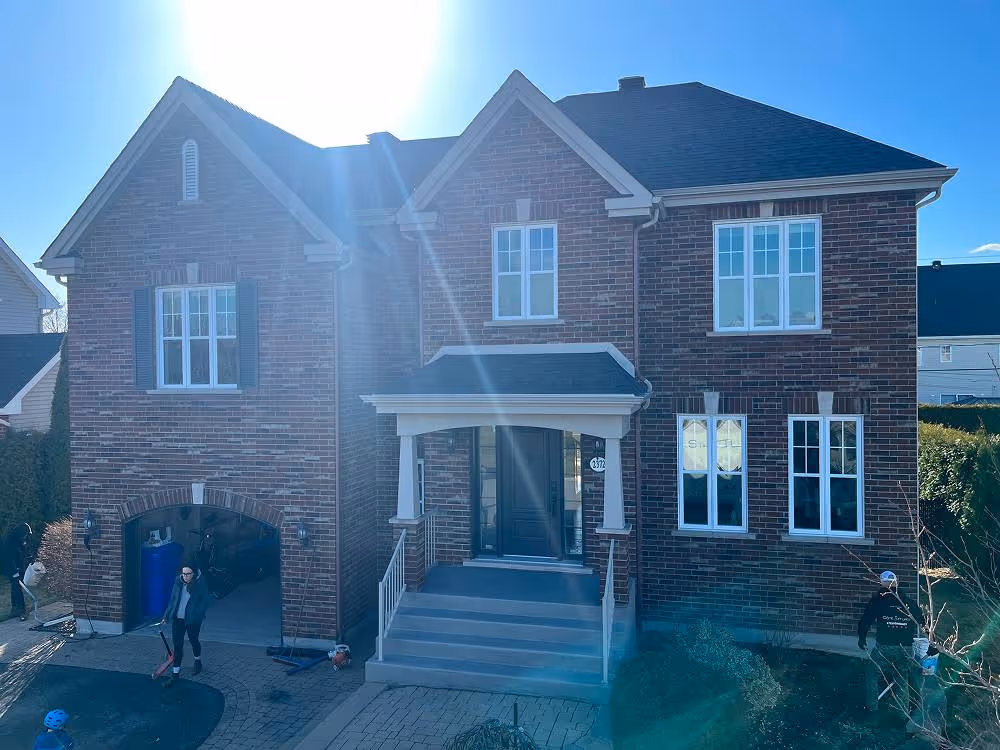 Maison en brique à deux étages avec une entrée principale avec porche, garage ouvert, plusieurs fenêtres blanches, et quelques personnes autour sous un ciel bleu ensoleillé.