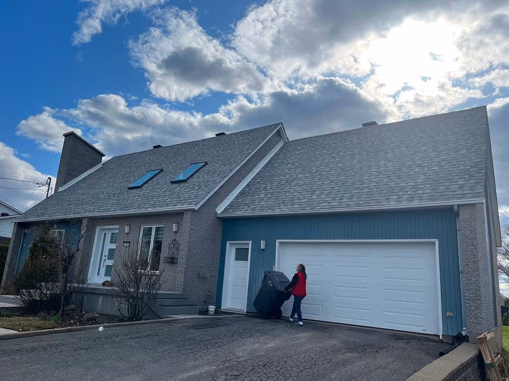Maison avec un garage double blanc, une porte d'entrée avec deux fenêtres de toit, et une personne en veste rouge déplaçant une poubelle noire sur l'allée.