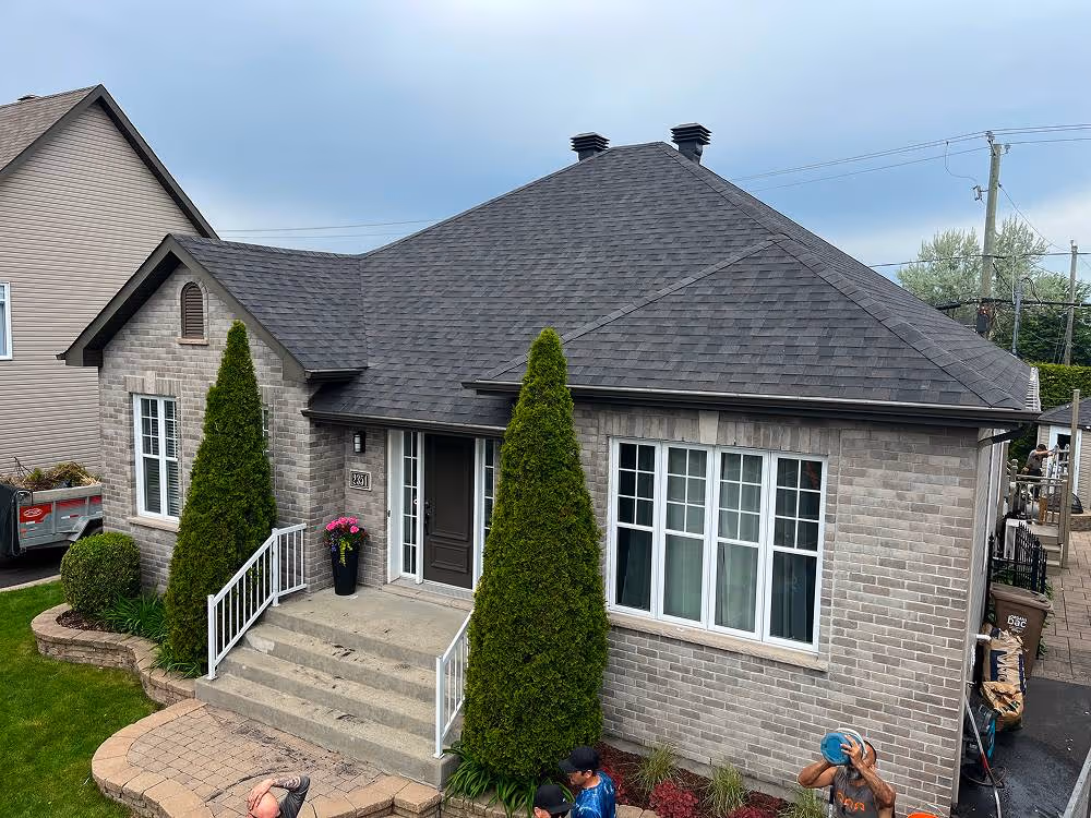 Maison de plain-pied en brique grise avec un toit en bardeaux noirs, deux grands sapins devant l'entrée et un petit jardin soigné.