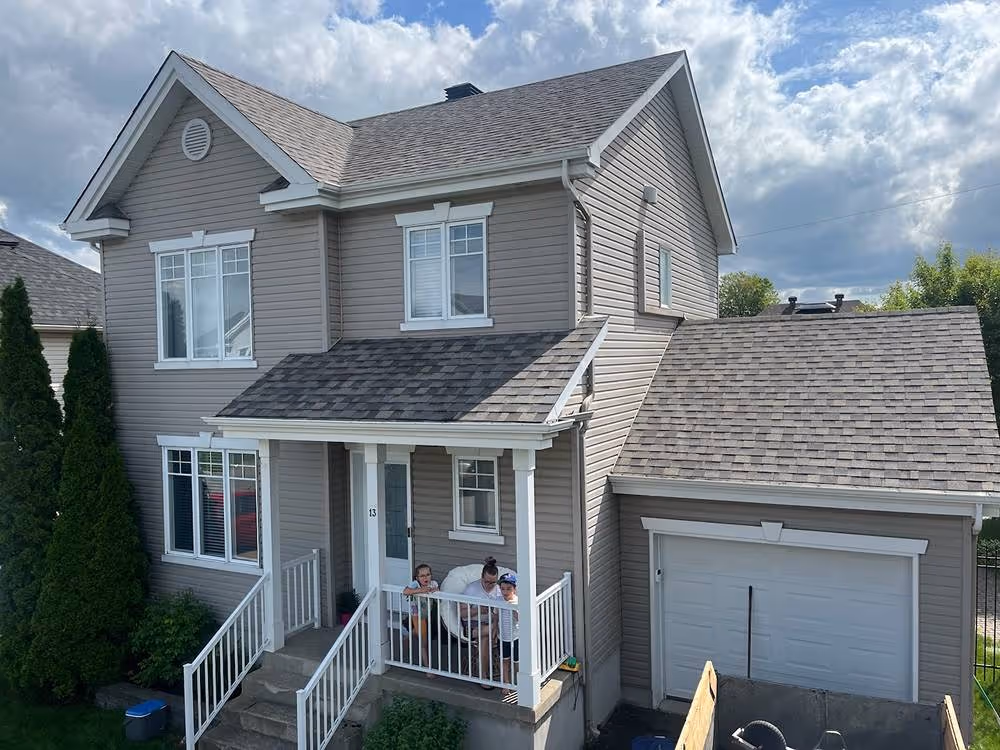 Maison grise à deux étages avec un porche blanc, deux enfants et un adulte assis sur le porche, garage attenant et ciel partiellement nuageux.