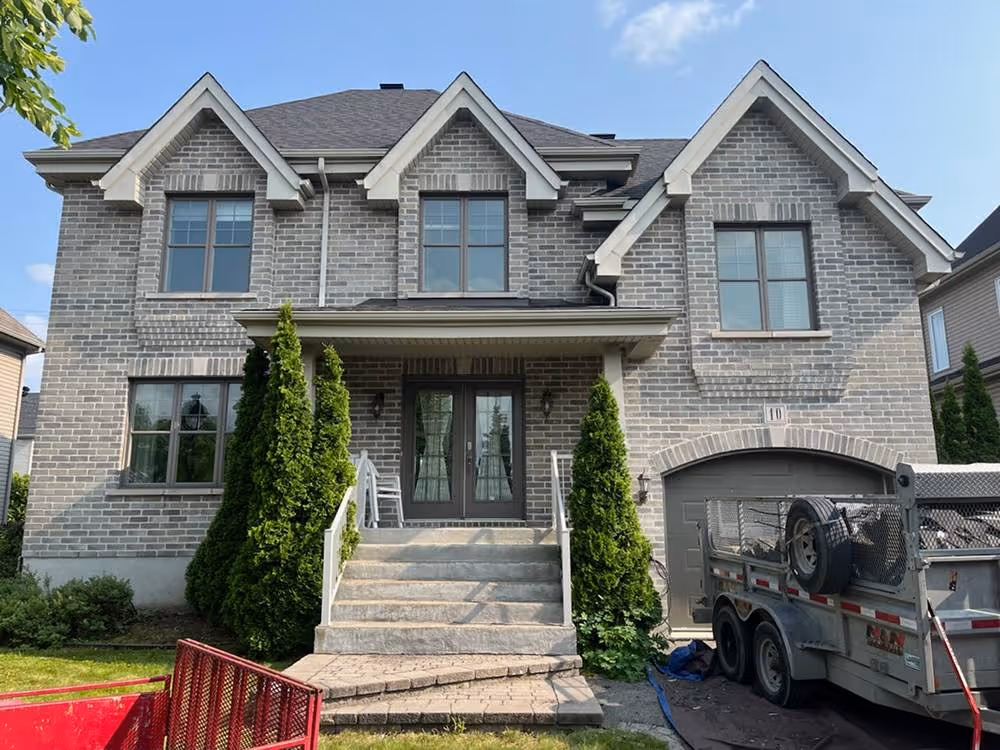 Vue aérienne de toits noirs en bardeaux sur plusieurs maisons en brique rouge entourées de végétation et voitures garées.