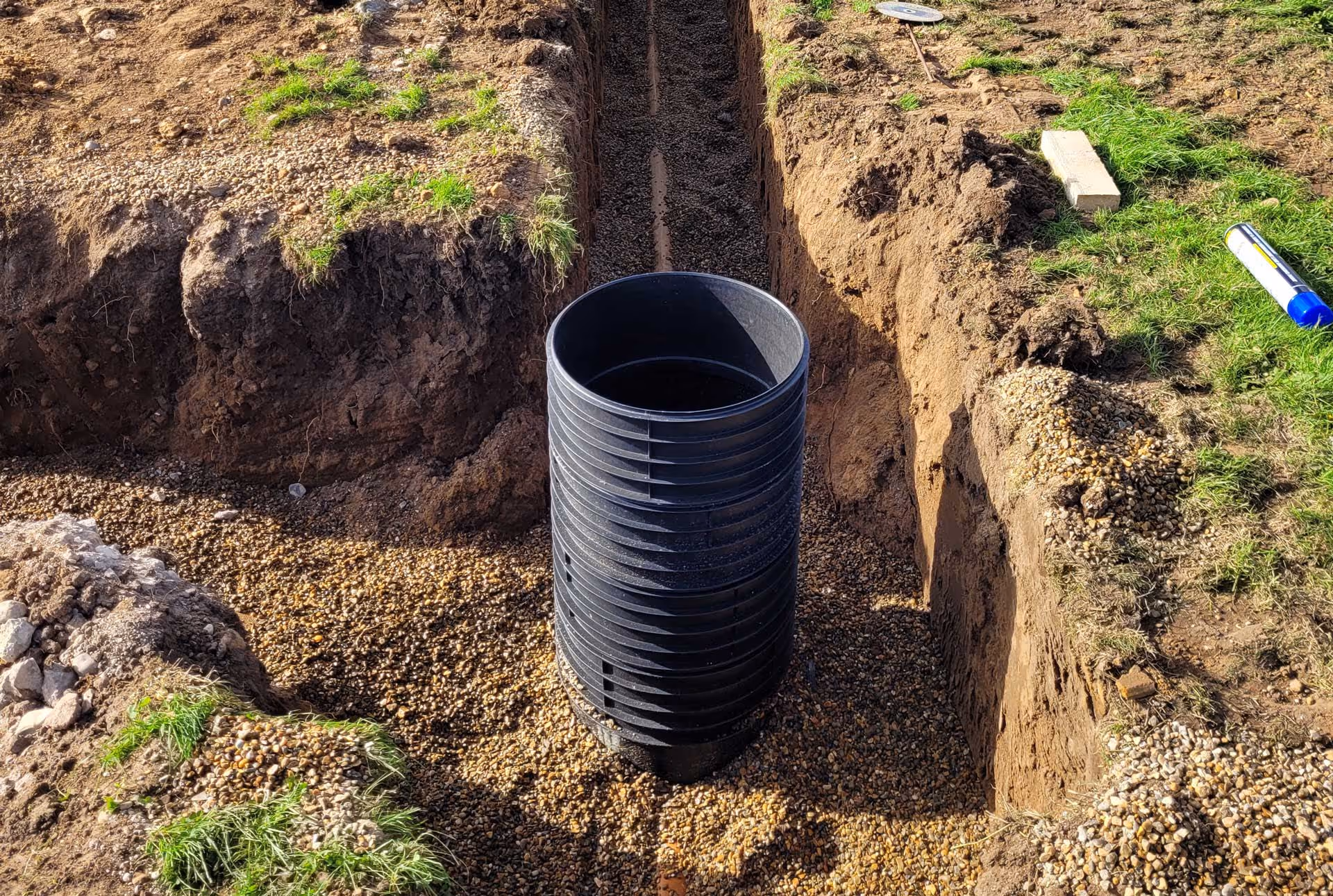 A large black drainage pipe is installed in a trench surrounded by dirt and gravel.