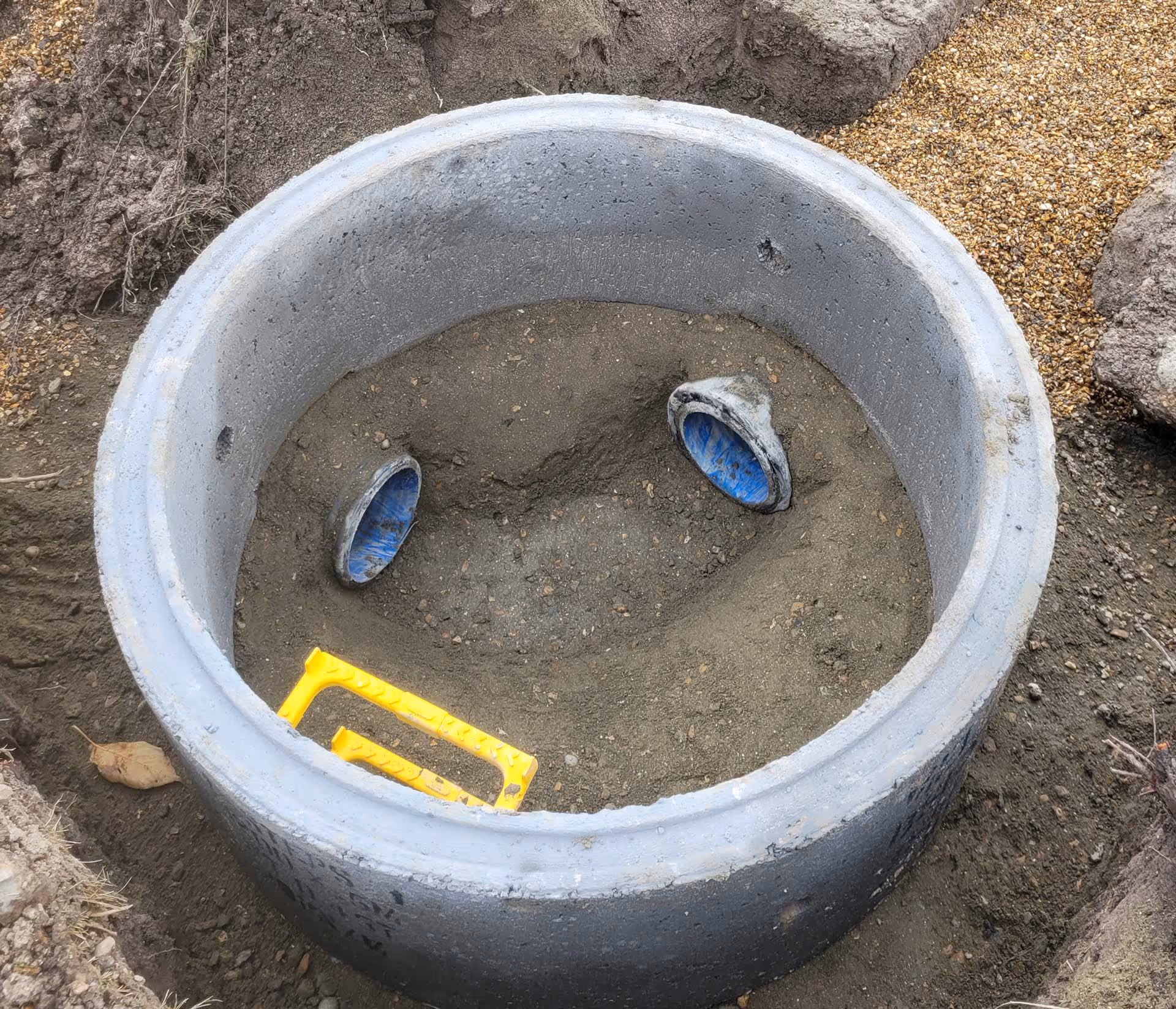 Concrete manhole with two blue pipes inside, partially filled with soil and surrounded by dirt.