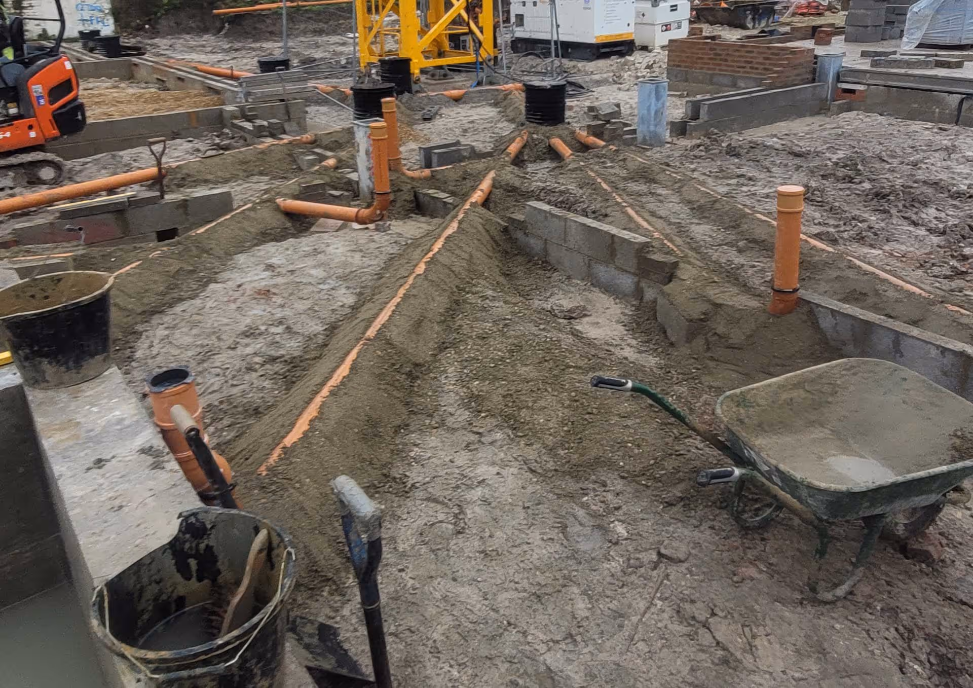 Construction site with exposed pipes and a wheelbarrow on a muddy ground.
