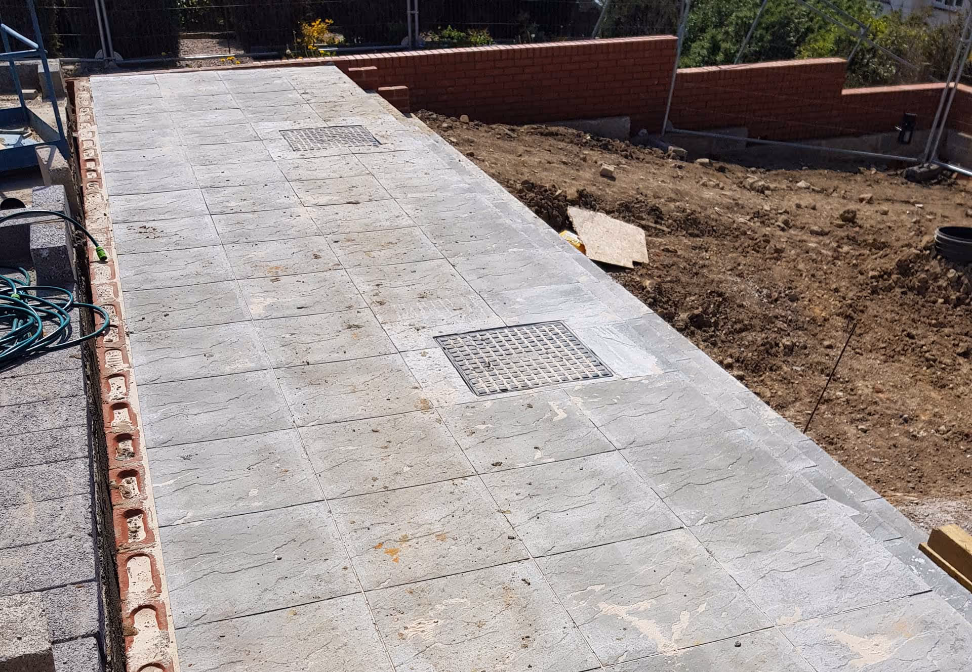 A newly paved walkway with concrete slabs and drainage grates sits next to a patch of soil.