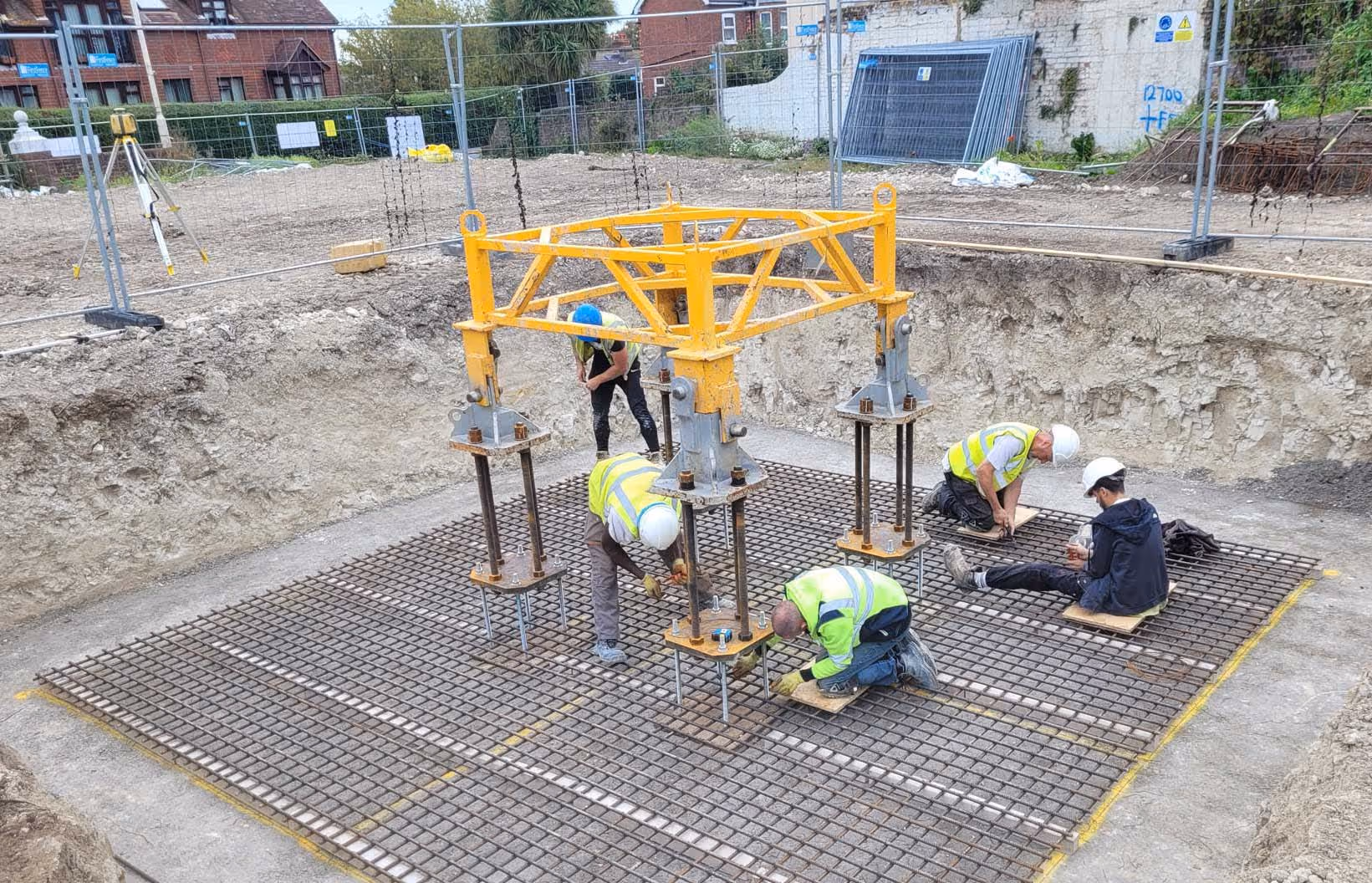 Five construction workers in safety gear install metal framework on a grid inside a large dug-out area.