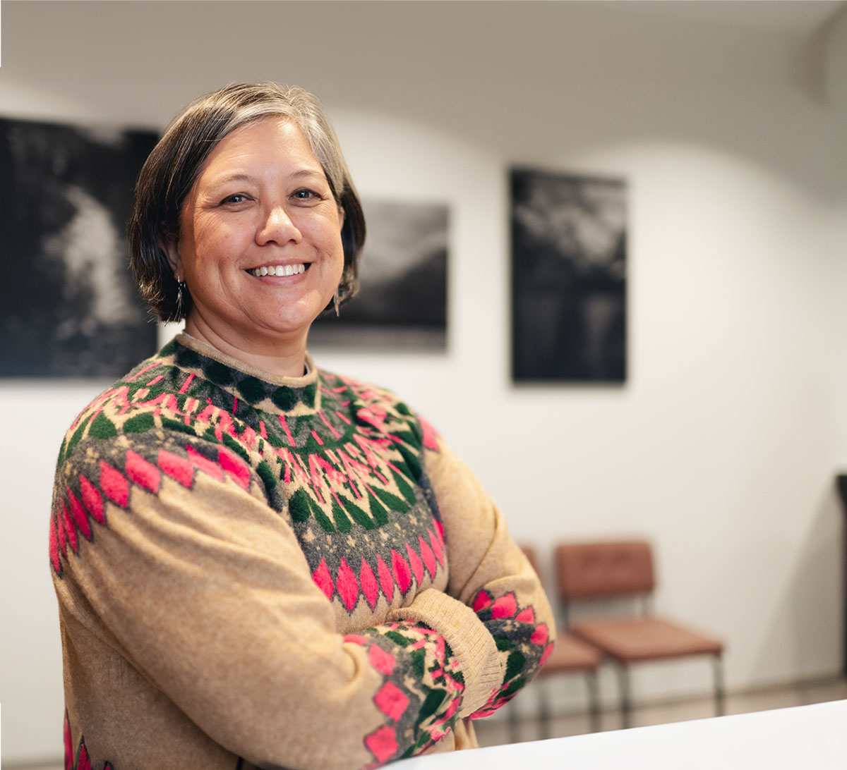 Smiling woman with short hair wearing a colorful patterned sweater standing with arms crossed in a room with blurred chairs and black-and-white wall art.