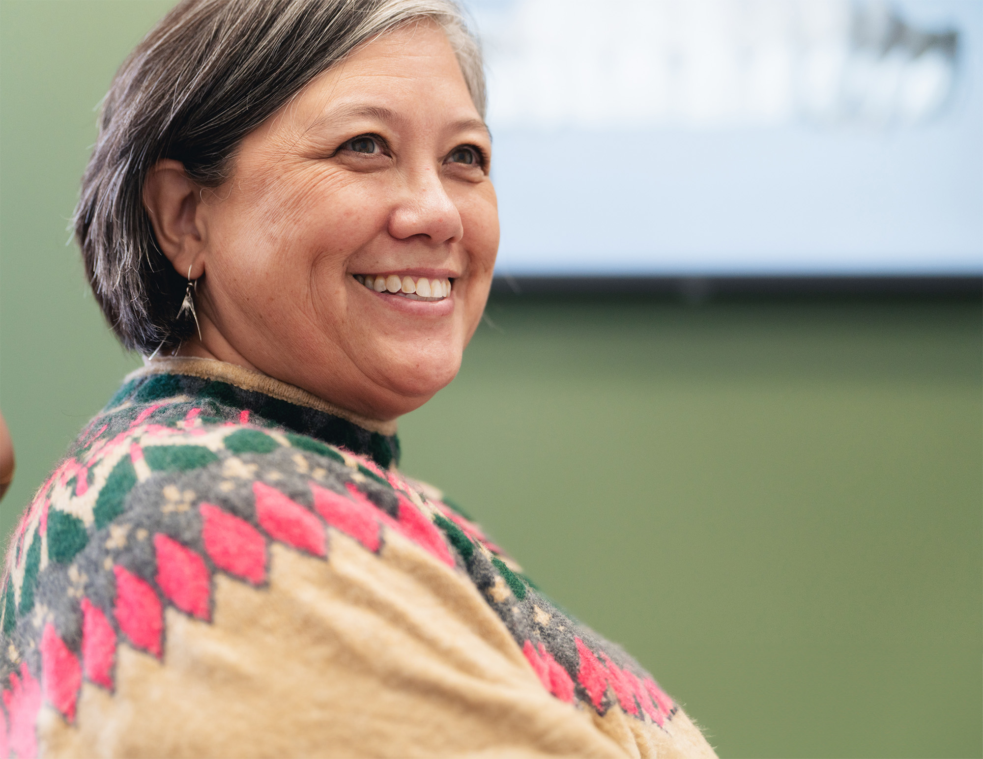 Smiling middle-aged woman with short gray hair wearing a colorful patterned sweater.