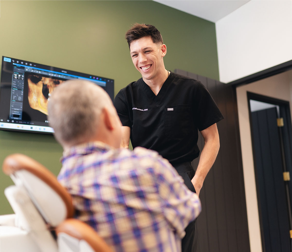 Smiling dentist in black scrubs shaking hands with an older patient seated in a dental chair.