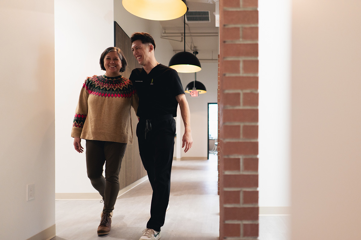Smiling woman in a patterned sweater walking arm-in-arm with a man in black scrubs in a well-lit hallway.