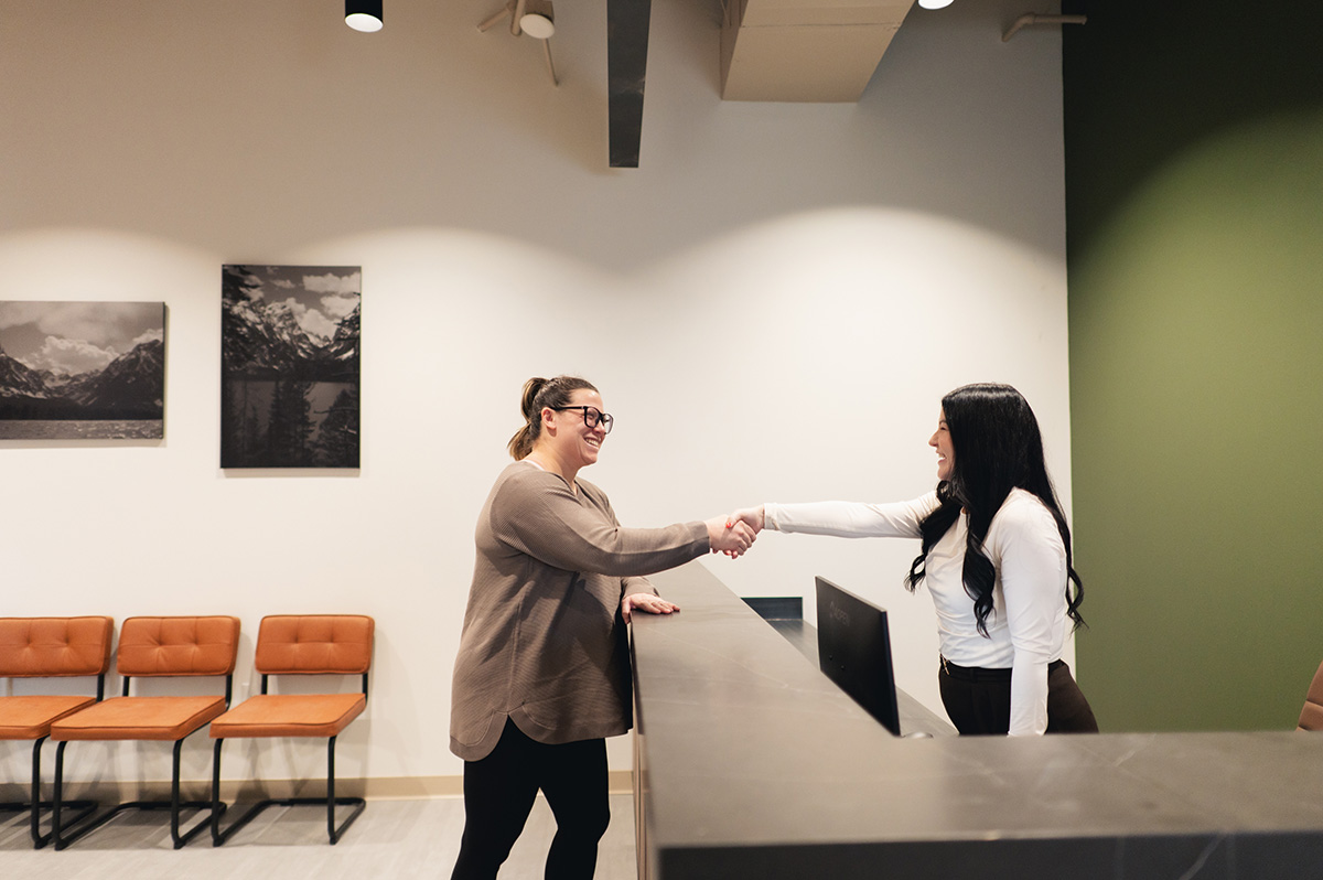 Two women smiling and shaking hands across a reception desk in a modern office.