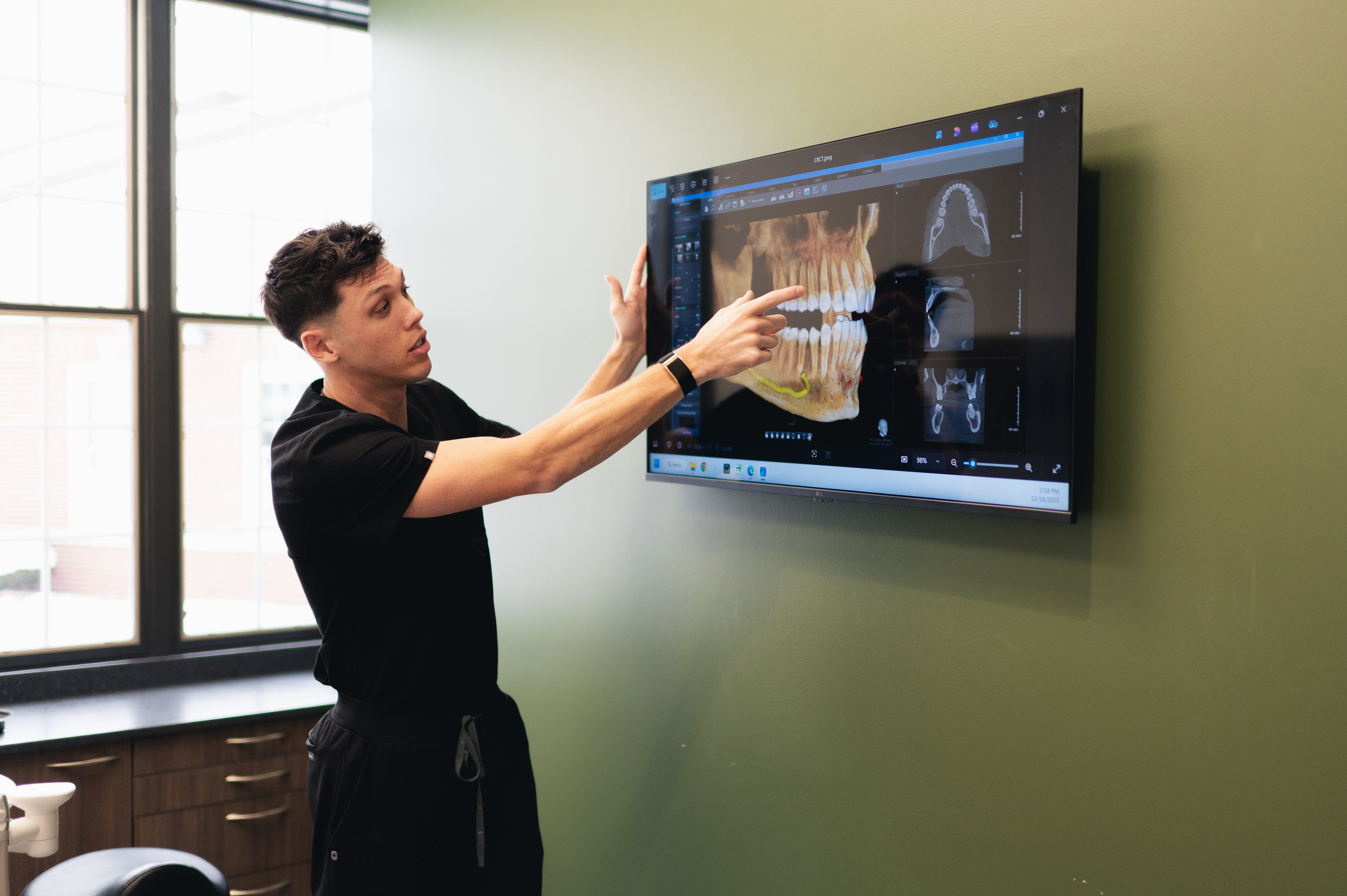 Dentist in black scrubs pointing to a dental X-ray on a wall-mounted screen in a clinic.