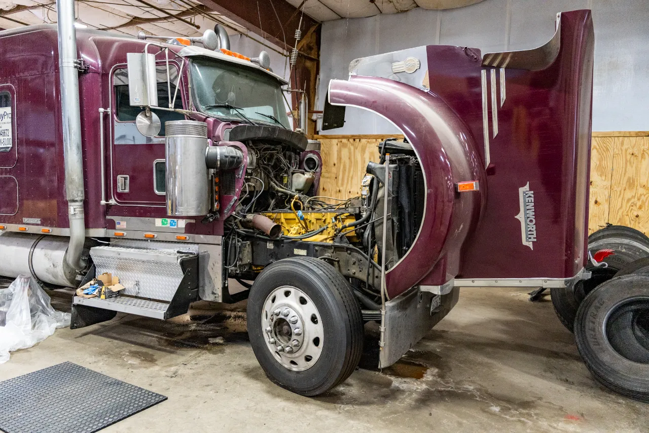 Kenworth truck undergoing engine repair with hood raised and exposed components in a maintenance shop.