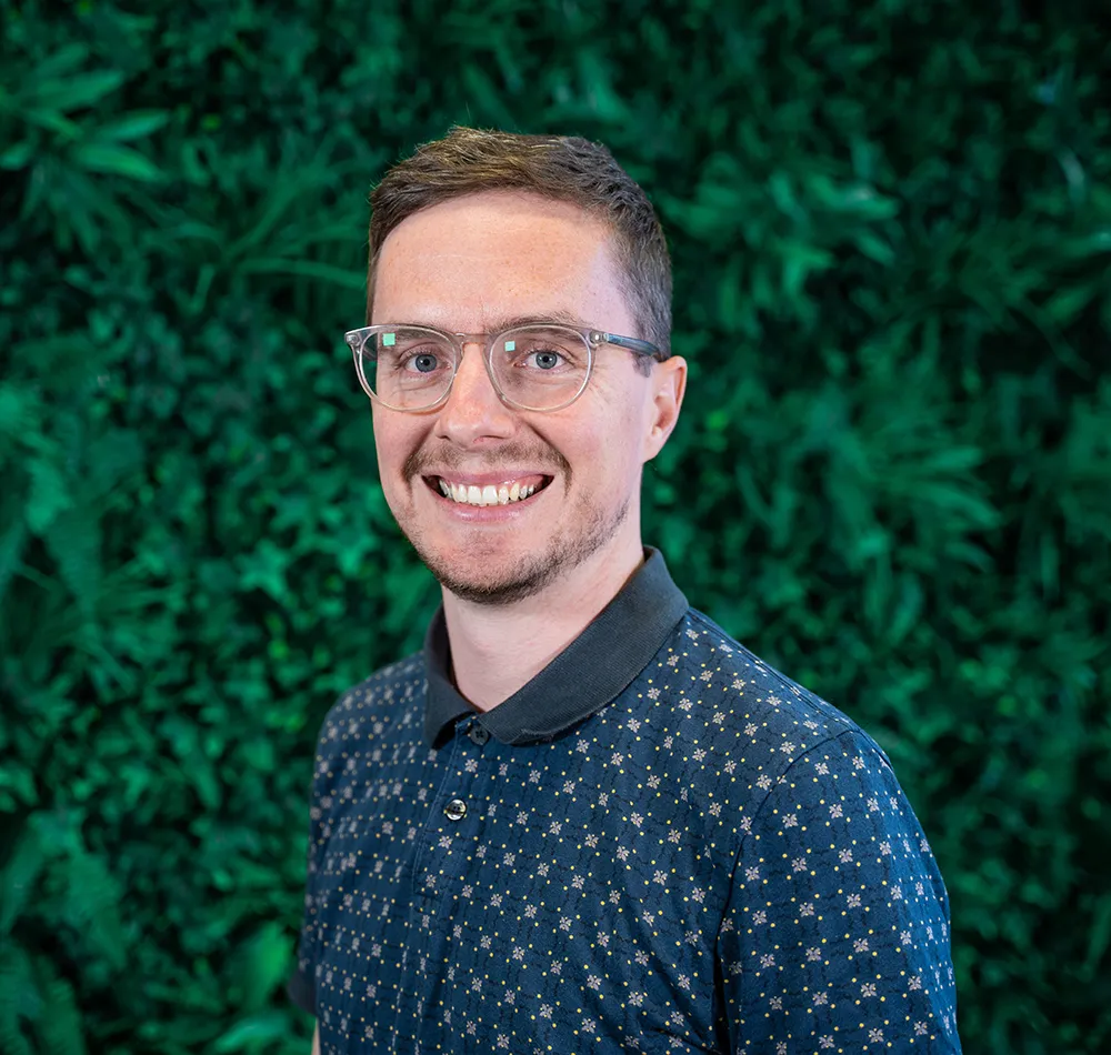 Smiling man with glasses in patterned shirt against green leafy background