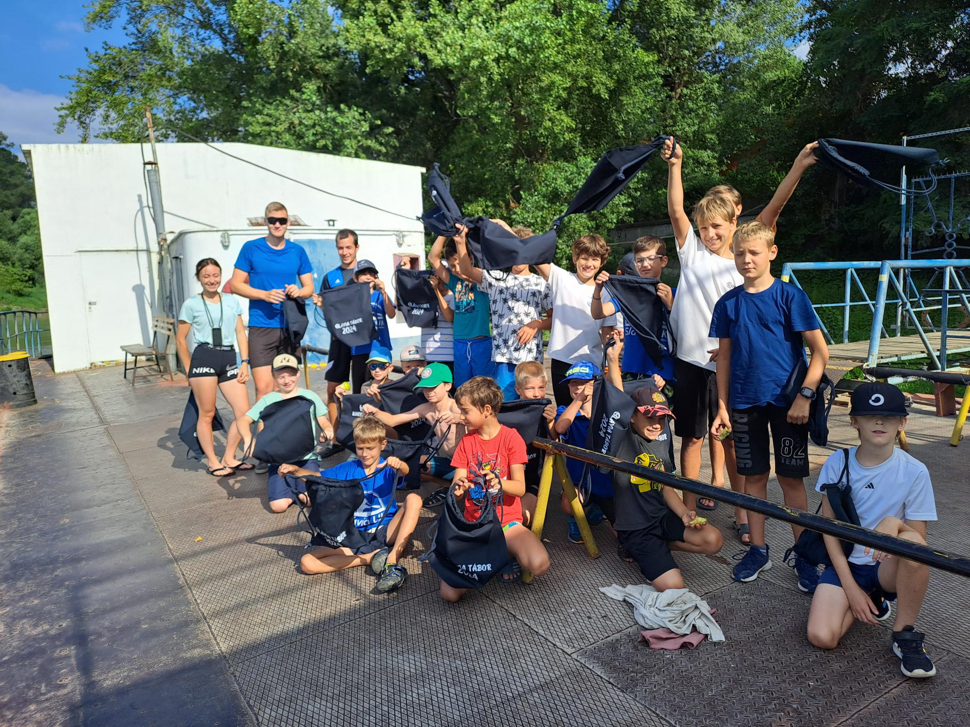 Group of children and two adults outdoors holding navy blue bags, posing on a metal platform with trees and a white building in the background.