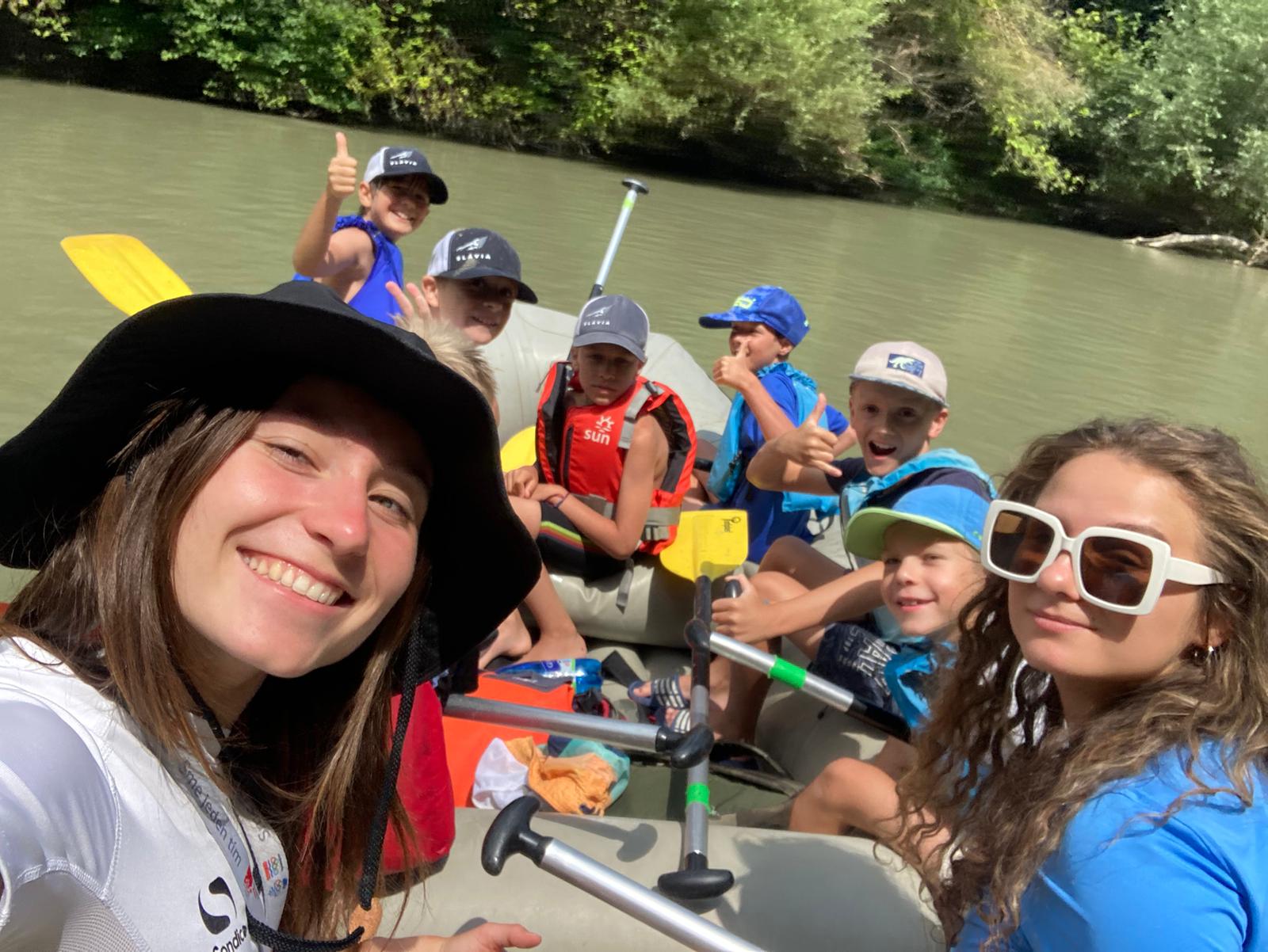 A group of children and two adults smiling on a raft in a calm river surrounded by green trees.