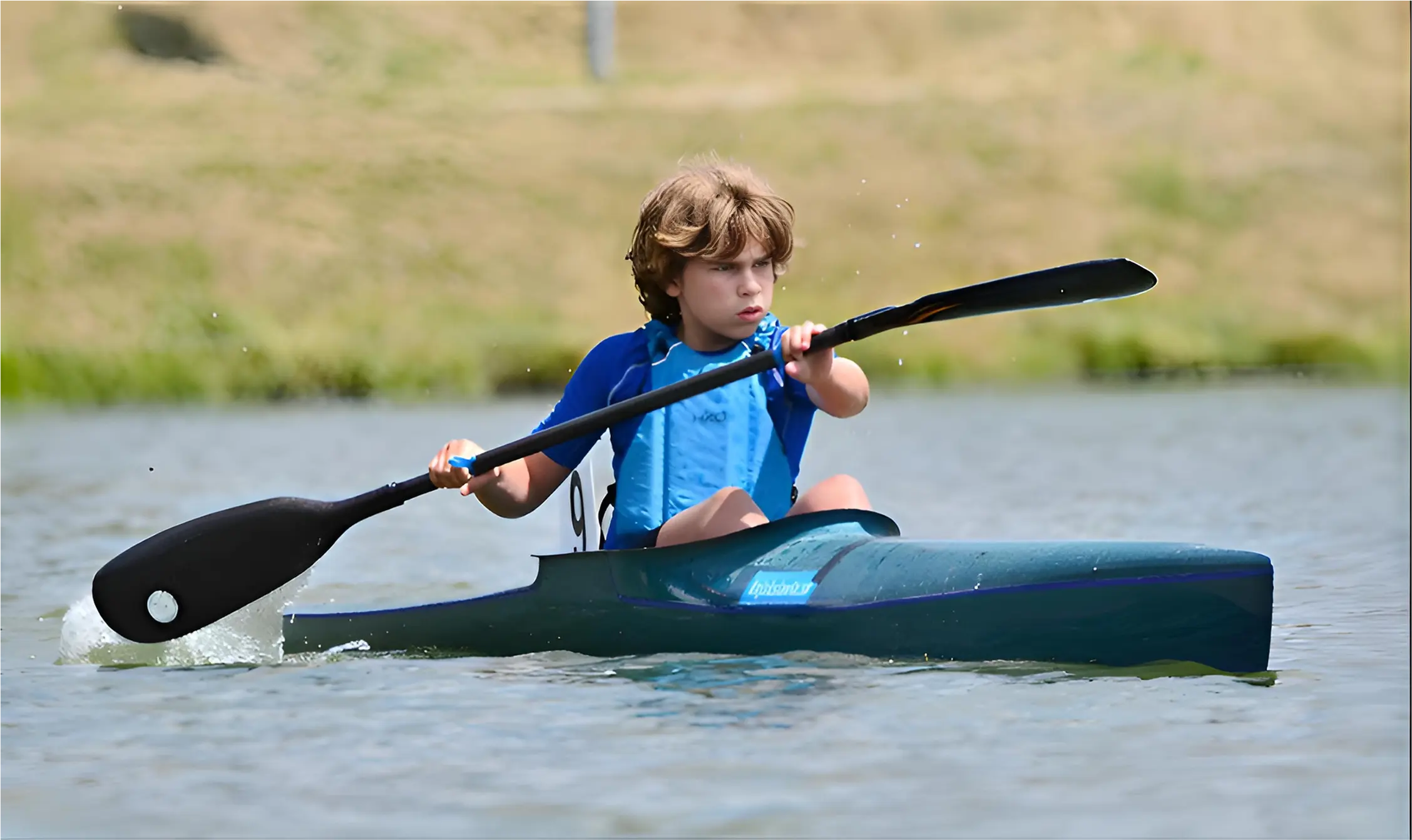 Young boy in blue shirt paddling a kayak on calm water with grassy background.