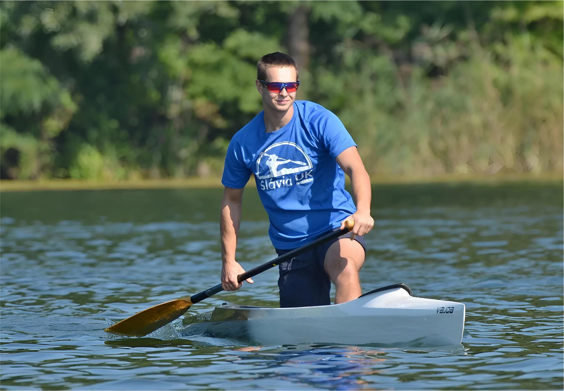 Man wearing sunglasses and a blue Slavia UK t-shirt paddling a white kayak on calm water with green foliage in the background.