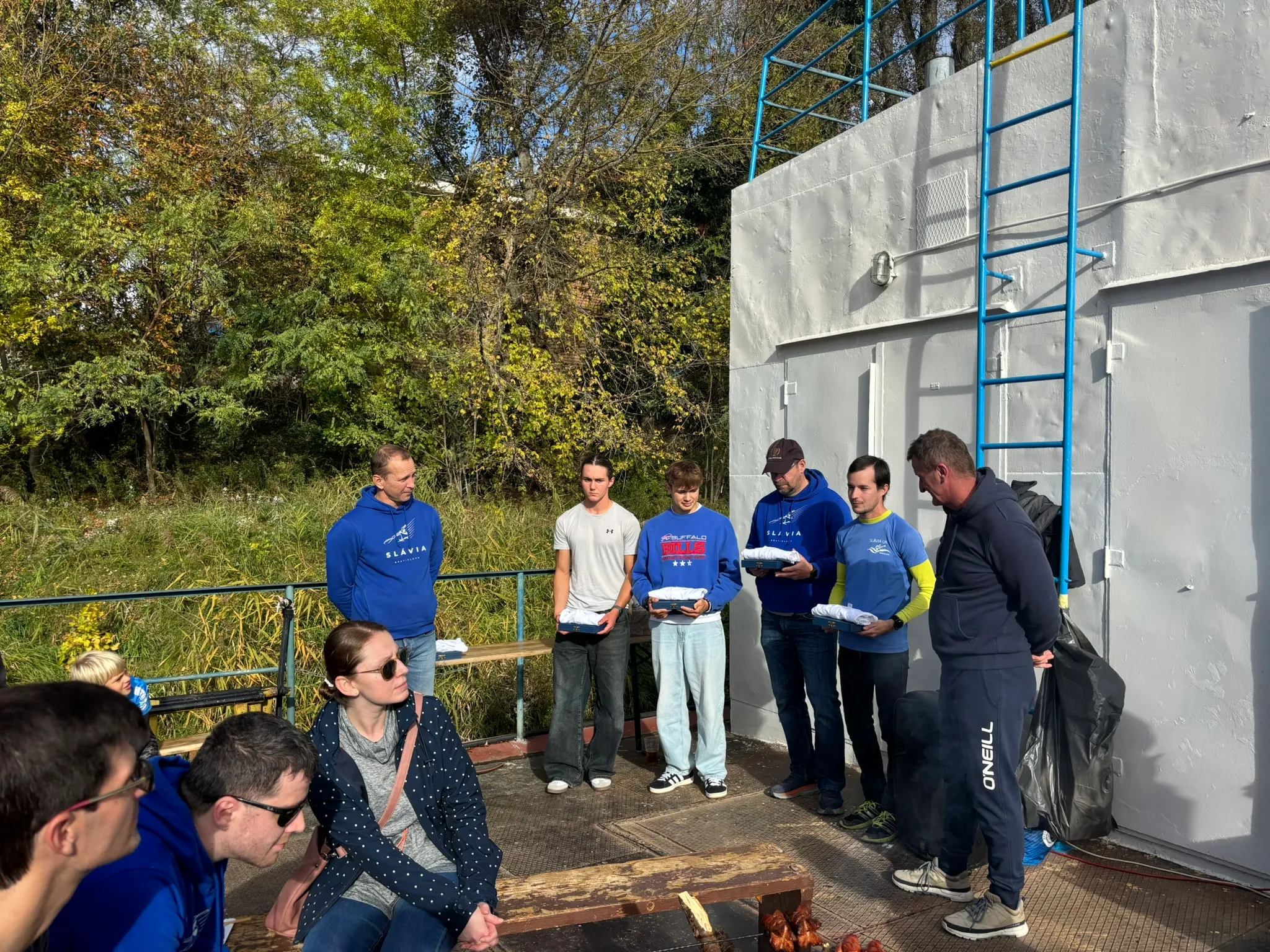 A group of people standing and sitting outdoors near a white wall with a blue ladder, some holding folded clothes, with trees in the background.