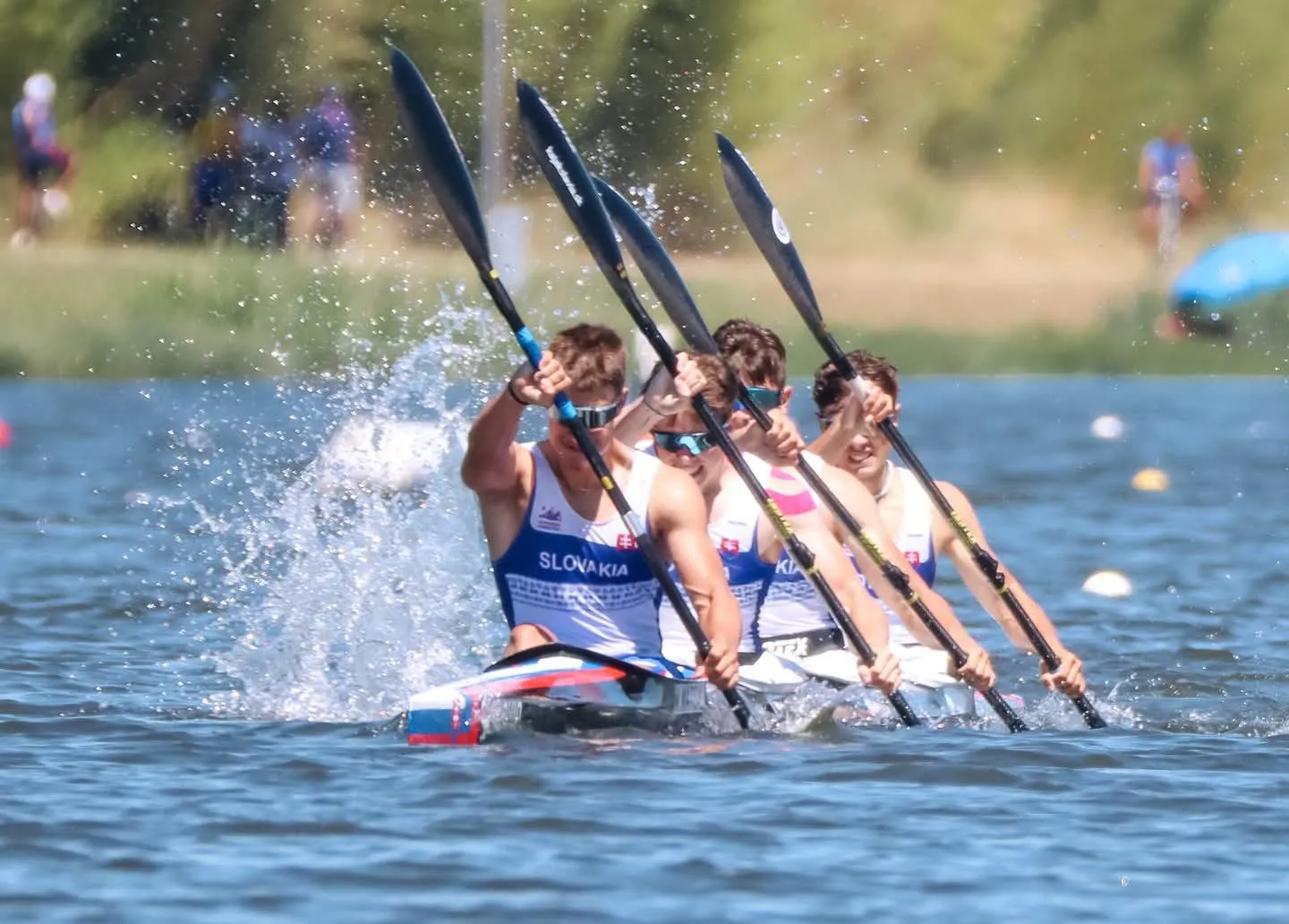 Four male athletes in a kayak race paddling strongly with water splashing around them, wearing Slovakia team uniforms.