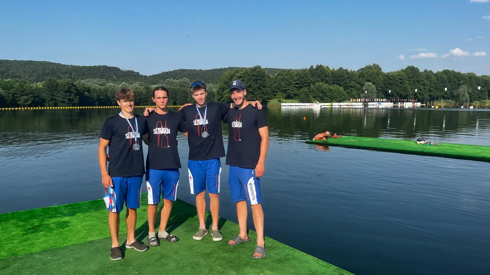 Four young men wearing Slovakia shirts and medals standing on a green dock by a calm lake with forested hills in the background.