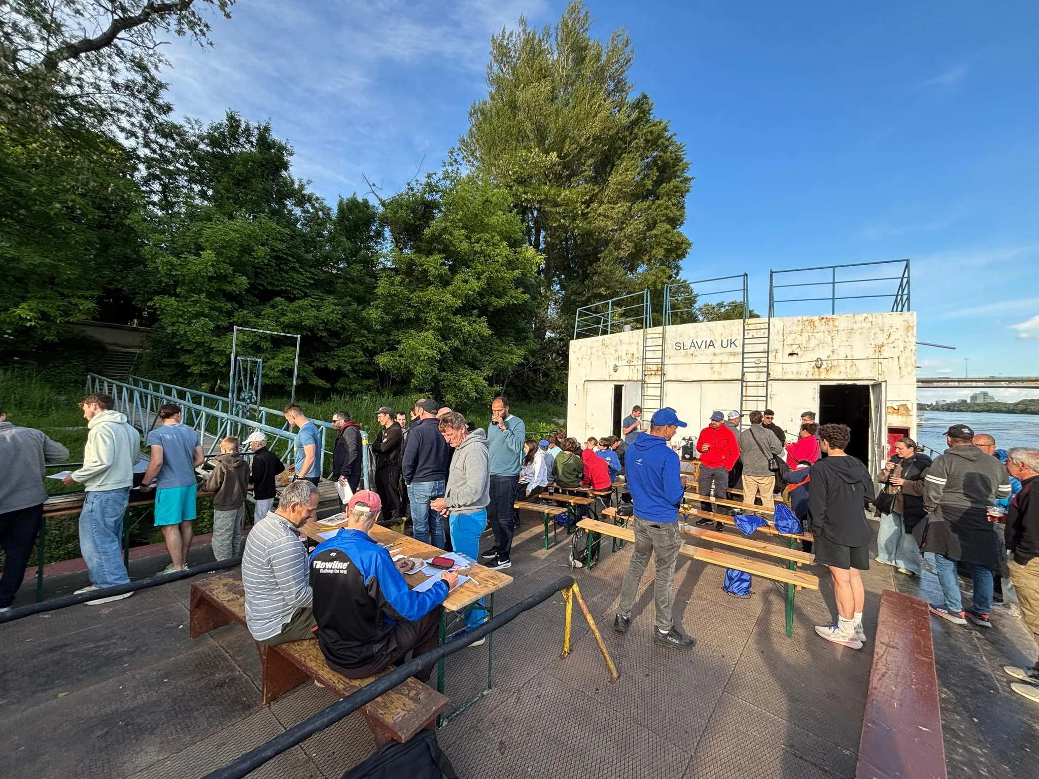 Group of people gathered outdoors near a river by a white building with 'SLÁVIA UK' signage, some seated at wooden benches and others standing in line.