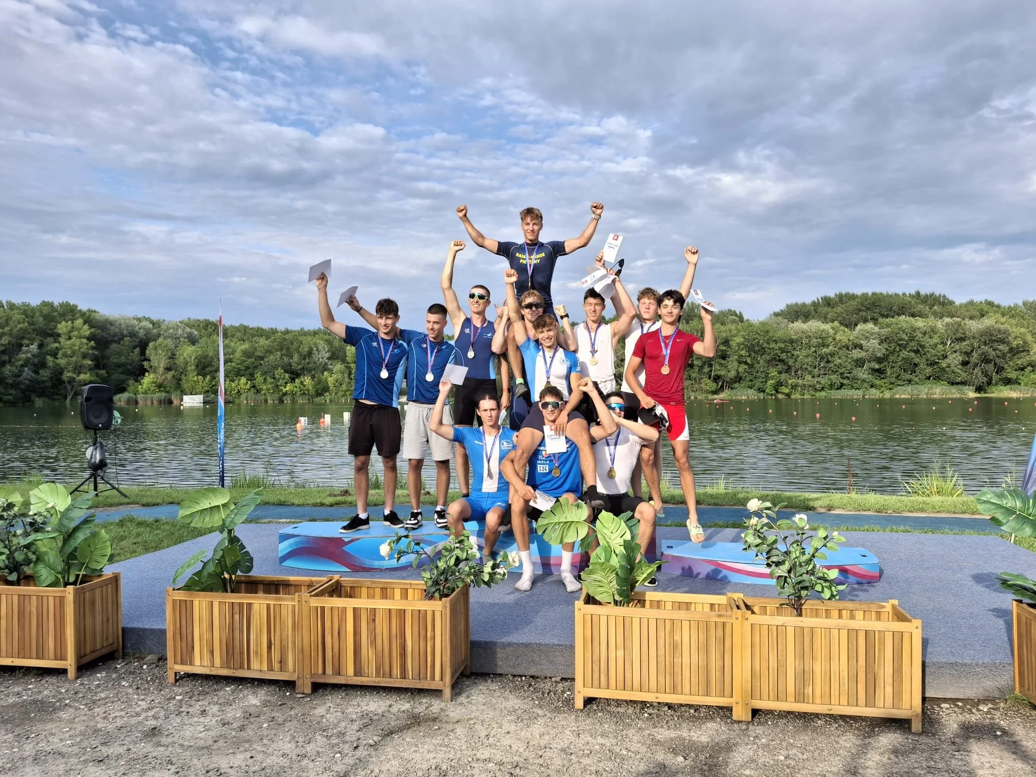 Group of young male athletes celebrating on a podium with medals and certificates at a lakeside outdoor event under a partly cloudy sky.