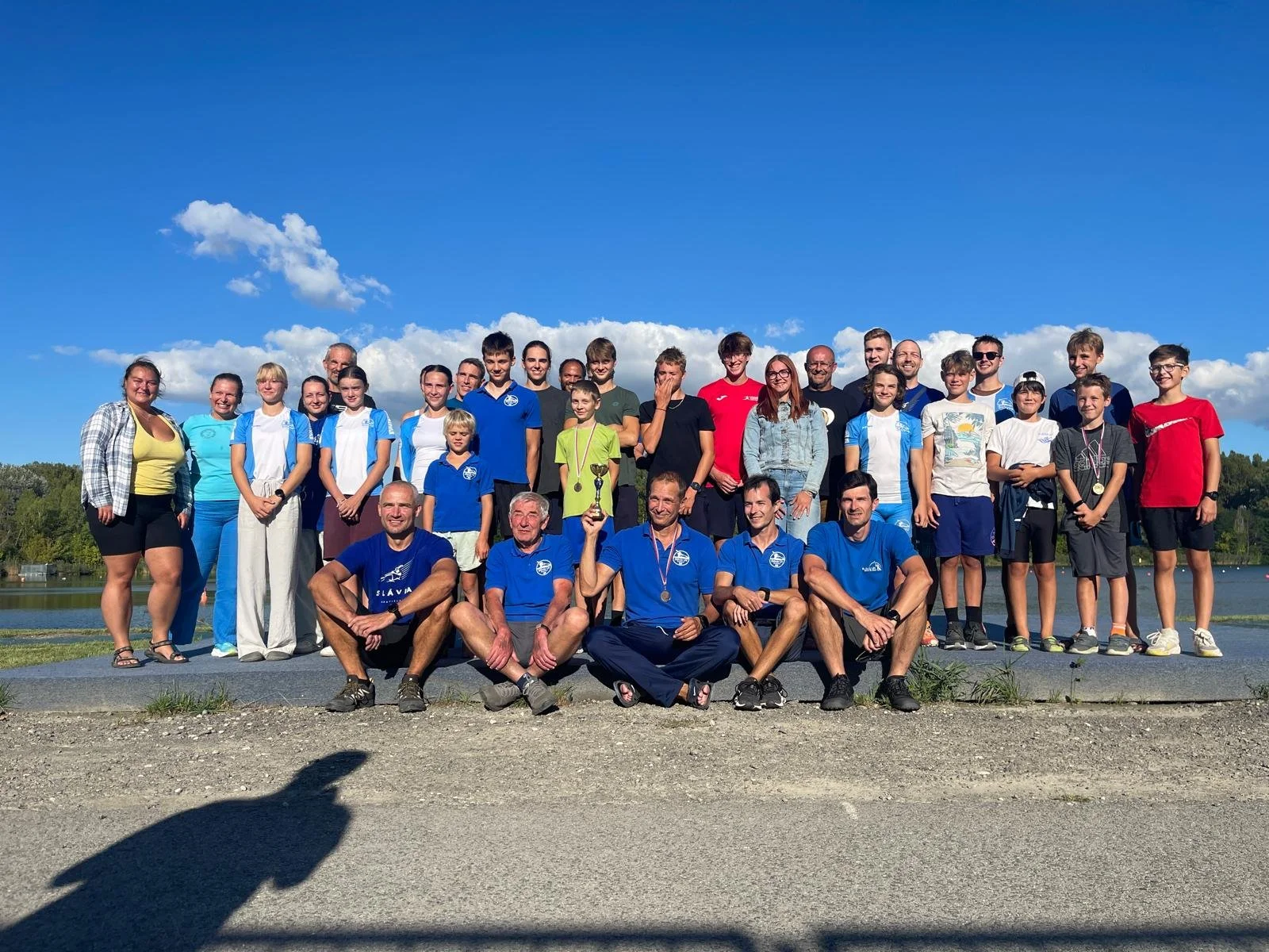 Large group of people of various ages posing outdoors near water under a blue sky, with one person holding a trophy and several wearing medals.