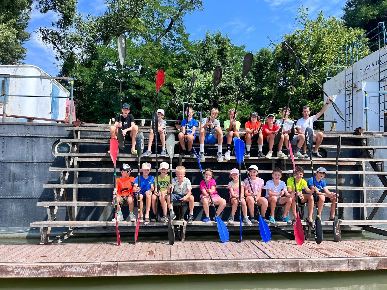 Group of children sitting on wooden bleachers holding kayak paddles under a clear blue sky.
