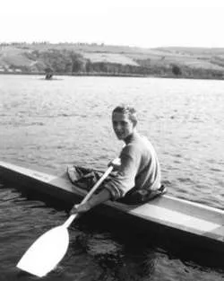 Black and white photo of a person sitting in a kayak on calm water holding a paddle, with a landscape of hills in the background.