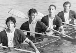 Four young men rowing together in a boat on the water, holding oars and wearing matching uniforms.