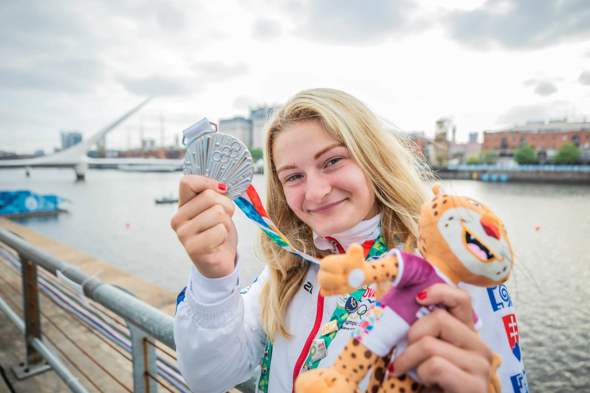 Smiling female athlete holding a silver Olympic medal and a spotted plush mascot by a waterfront with a modern bridge in the background.