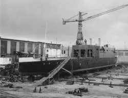 Black and white photo of a submarine on dry dock with a crane in the background and workers nearby.
