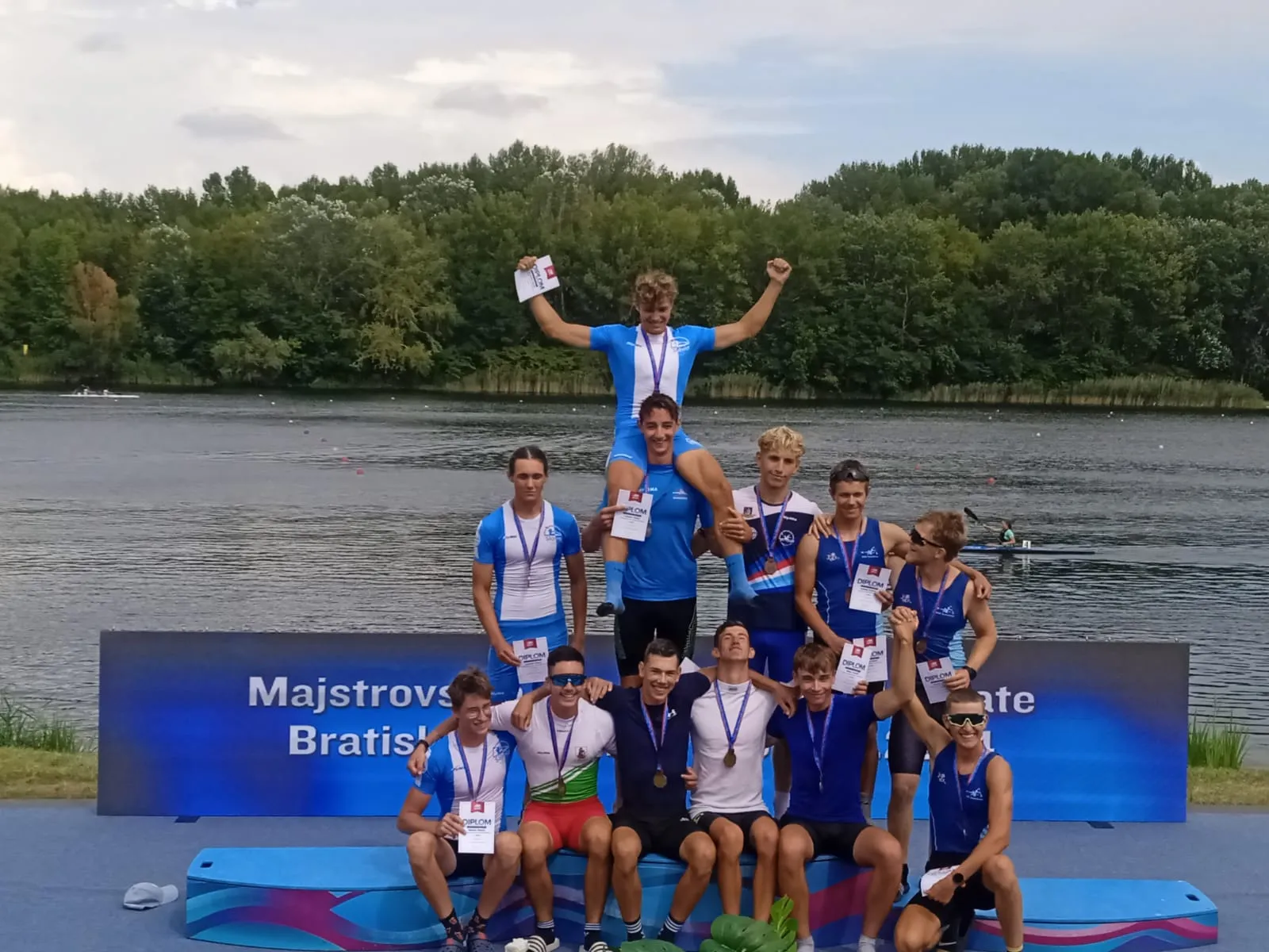 Group of young male rowers wearing medals and holding diplomas posing on a podium by a lake with trees in the background.