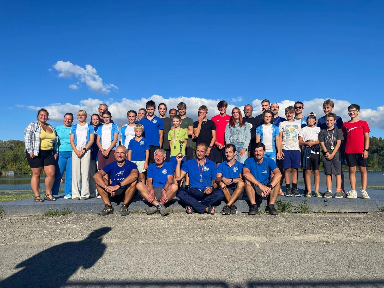 Group of smiling men, women, and children posing outdoors by a lake under a blue sky, some wearing medals and holding a trophy.