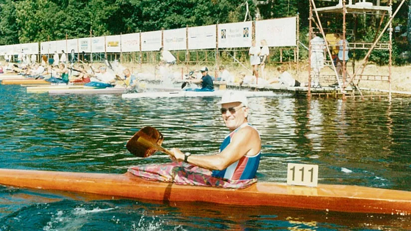 Smiling man in a white cap paddling a wooden kayak numbered 111 on calm water during a kayak race with other participants in the background.