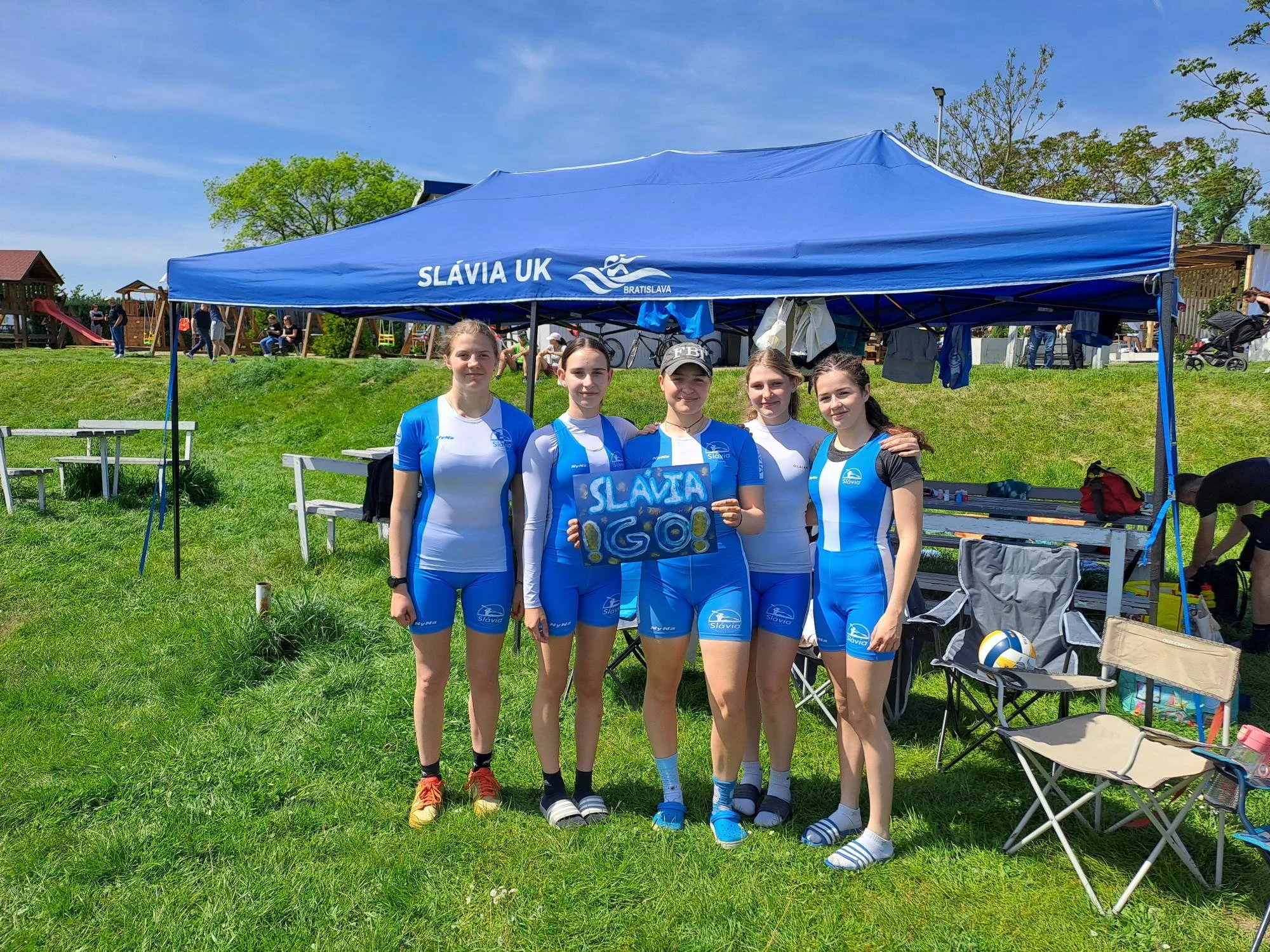 Five female athletes in blue and white Slávia UK cycling uniforms standing under a blue canopy tent on green grass, one holding a sign that says 'SLAVIA GO!'