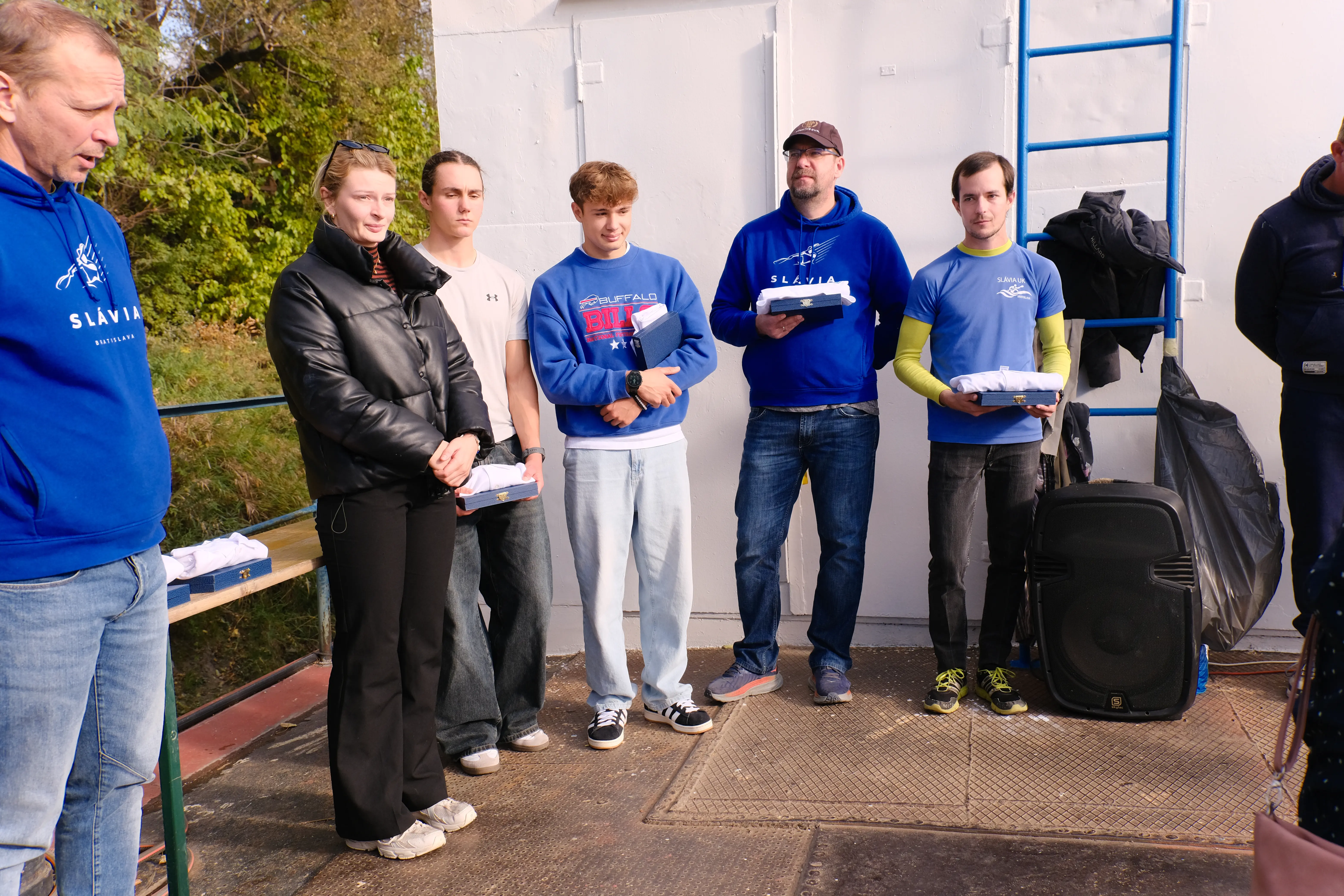 Group of six people standing outdoors, some holding folded clothes and boxes, wearing casual fall clothing including blue and black jackets and hoodies.