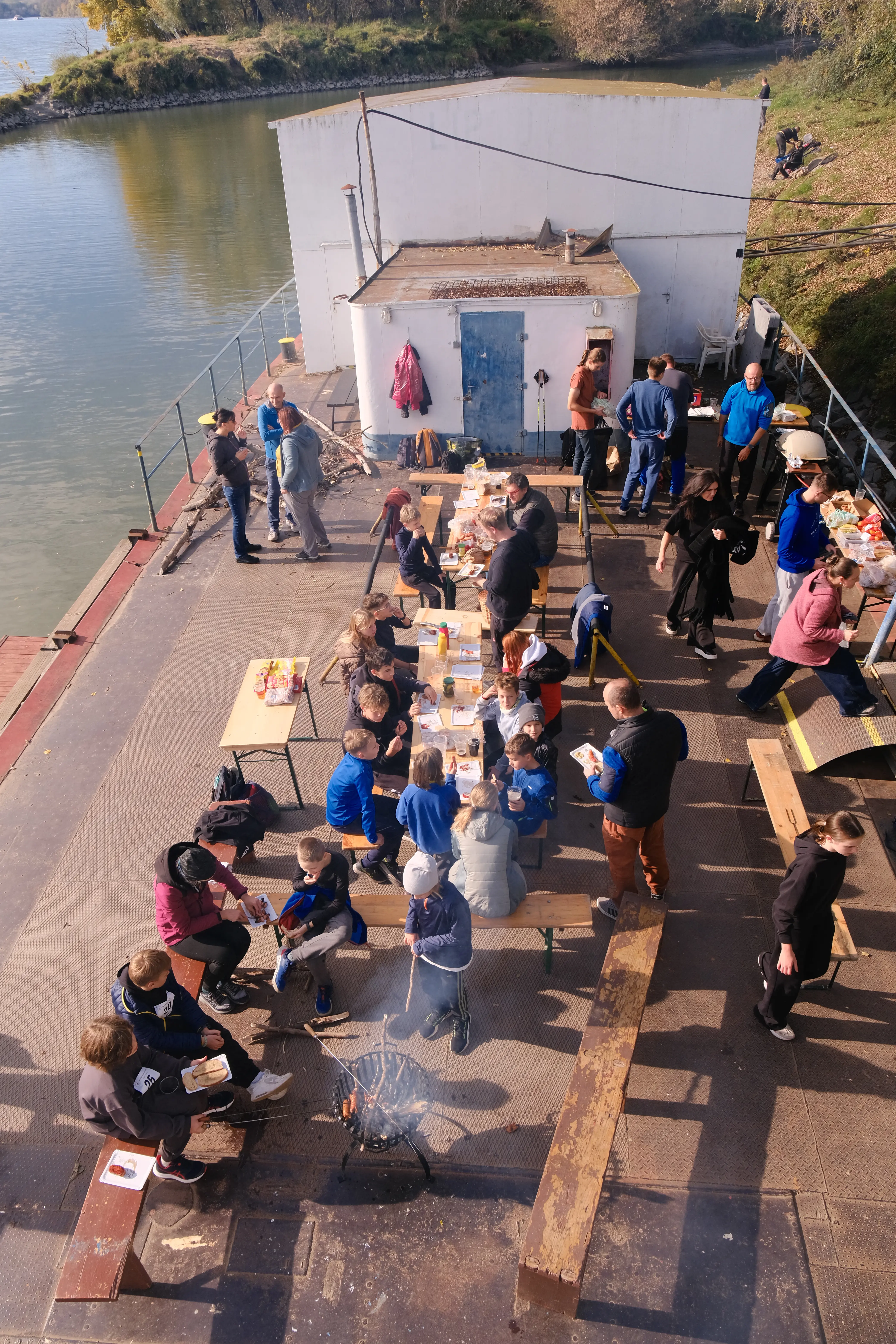 Group of people enjoying an outdoor barbecue by a river, sitting and standing around tables with food and drinks on a sunny day.