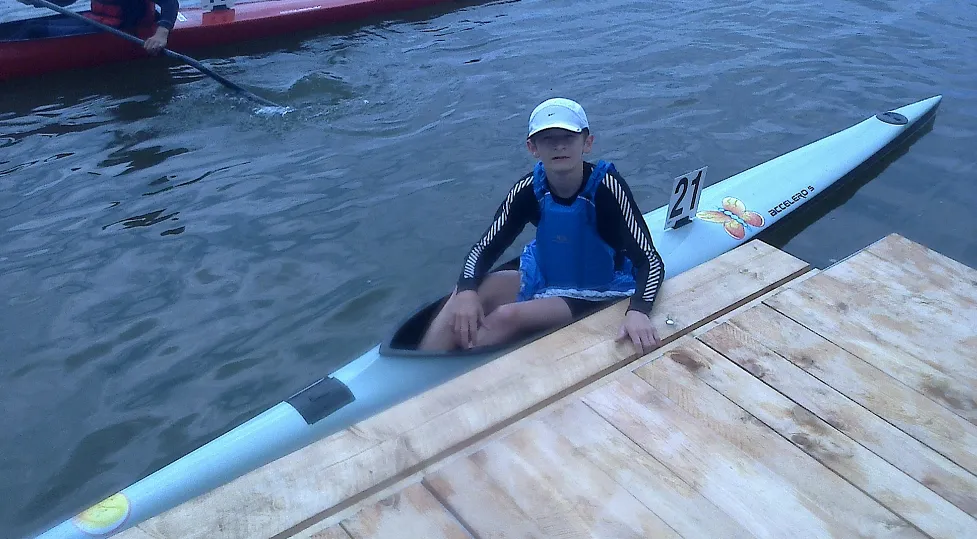 Young person wearing a white cap and blue life jacket sitting in a narrow white kayak numbered 21 at a wooden dock.
