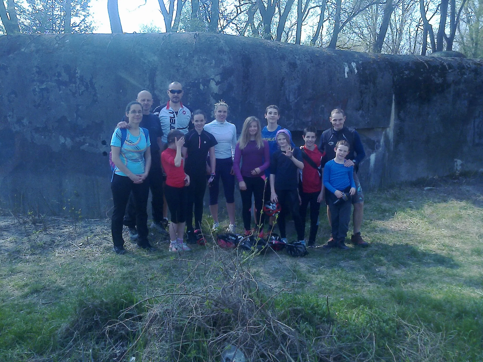 Group of eleven people, including adults and children, standing outdoors in front of a large concrete structure with trees in the background.